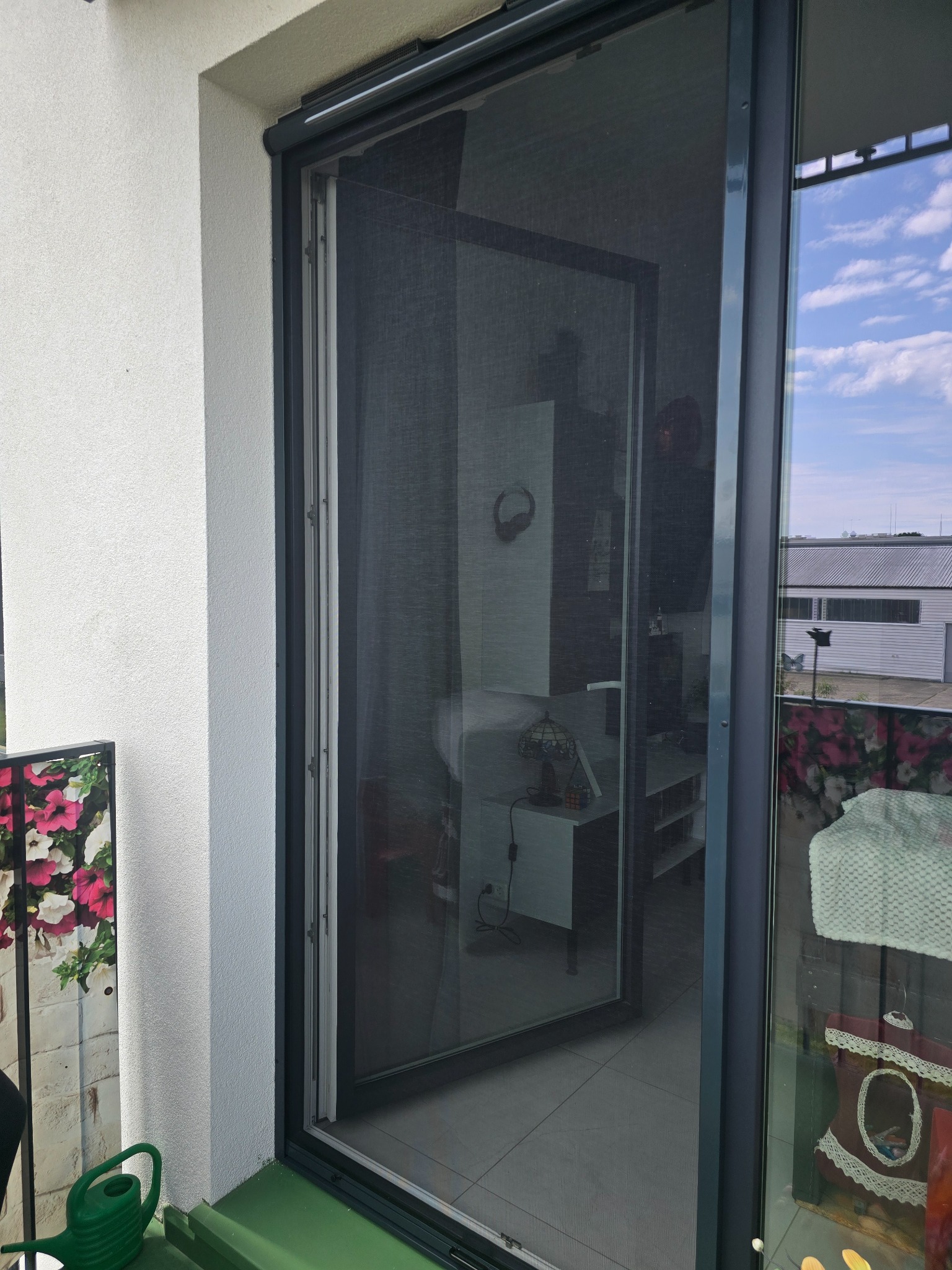 Zamontowana moskitiera ramkowa w nowoczesnym oknie balkonowym, widok z balkonu z zieloną podłogą i dekoracjami kwiatowymi, zarys wnętrza mieszkania w tle.