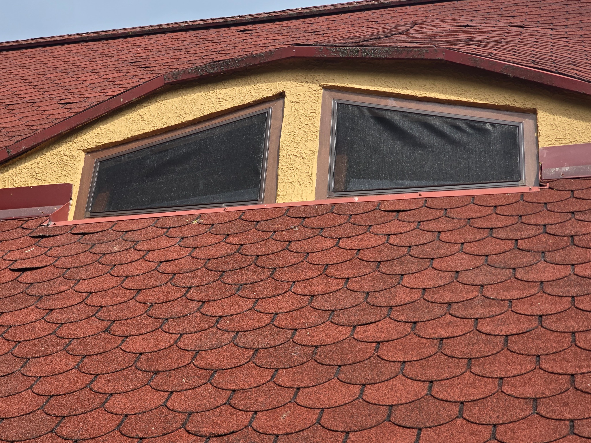 Dwa okna dachowe z moskitierami w żółtej ścianie na tle czerwonego dachu z gontu bitumicznego. Ujęcie z dołu, widok na zamontowane siatki.
