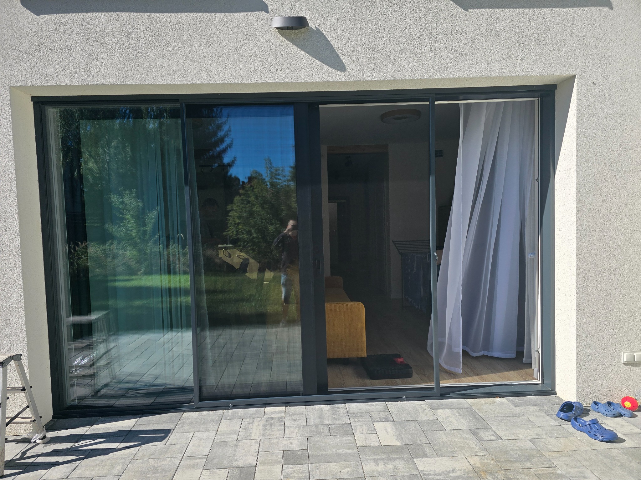 Przesuwne drzwi tarasowe z zamontowaną moskitierą, widok z zewnątrz. Na pierwszym planie szare płytki chodnikowe i drabina oparta o ścianę.