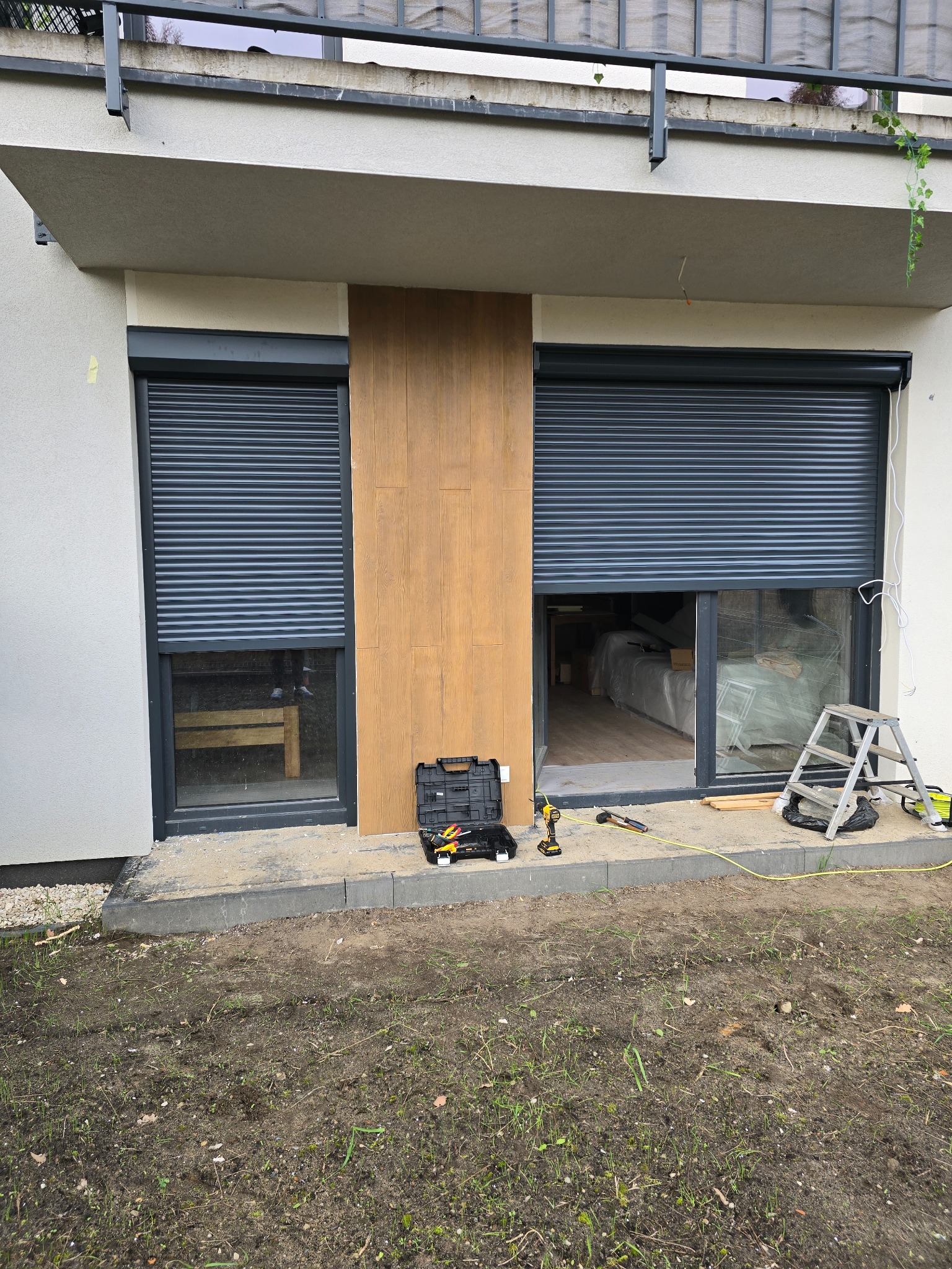 Montaż szarych rolet zewnętrznych w nowoczesnym domu. Widoczne narzędzia, drabina i częściowo opuszczona roleta nad oknem tarasowym.