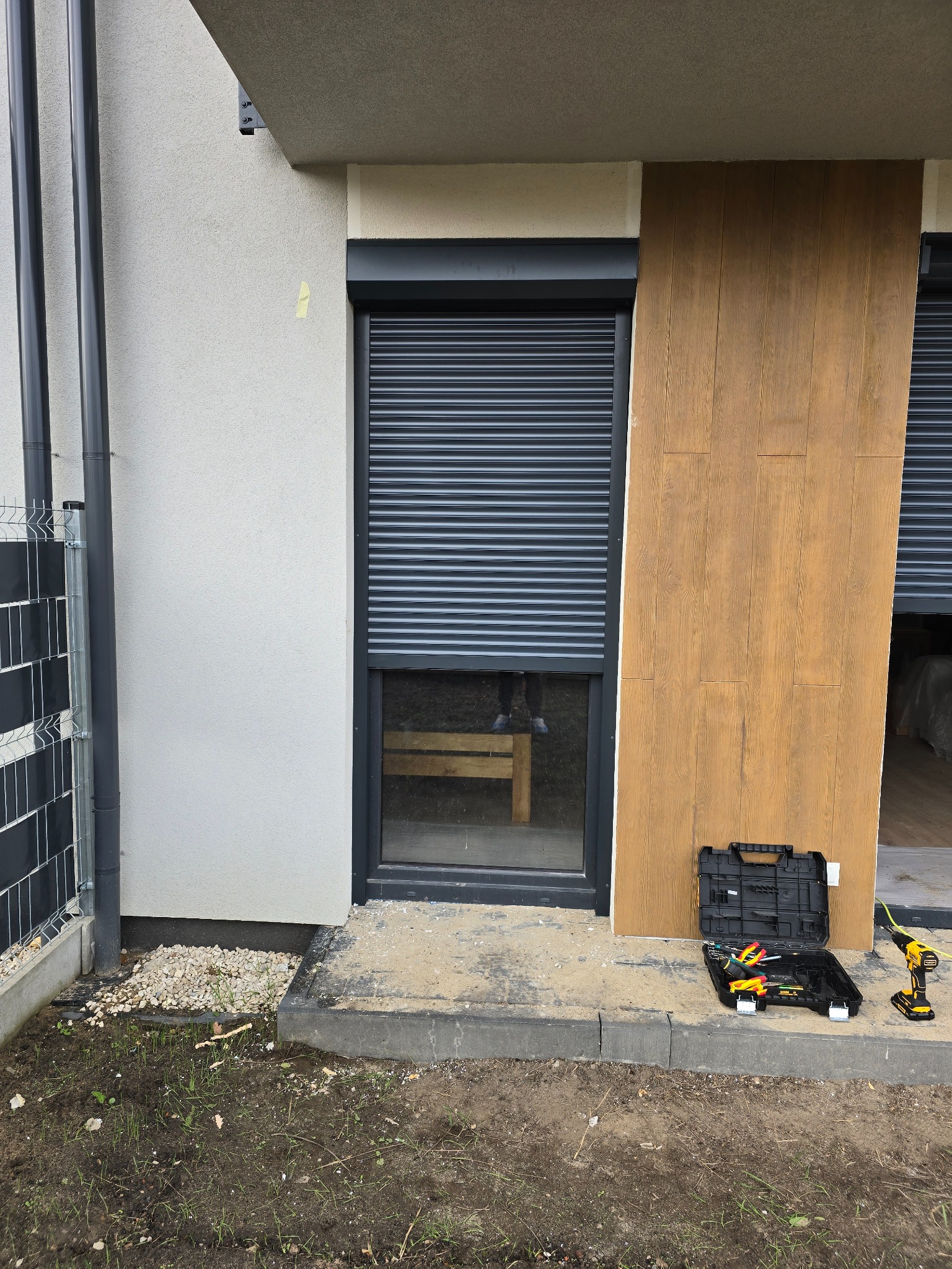 Zamknięta, grafitowa roleta zewnętrzna na oknie nowoczesnego domu. Obok skrzynka z narzędziami i wkrętarka. Widoczne elementy elewacji i fragment trawnika.