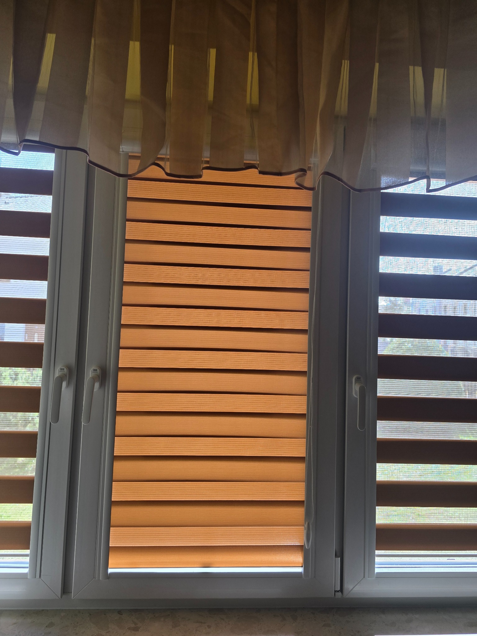 Okna z zamontowanymi, częściowo zasłoniętymi roletami dzień-noc w odcieniach brązu, z delikatną firanką w górnej części kadru. Widoczne uchwyty okienne.