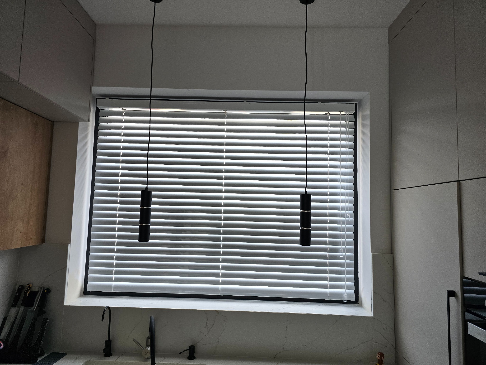 Białe żaluzje poziome w oknie kuchni z marmurowym blatem i nowoczesnymi lampami wiszącymi. Minimalistyczny design, jasne wnętrze.