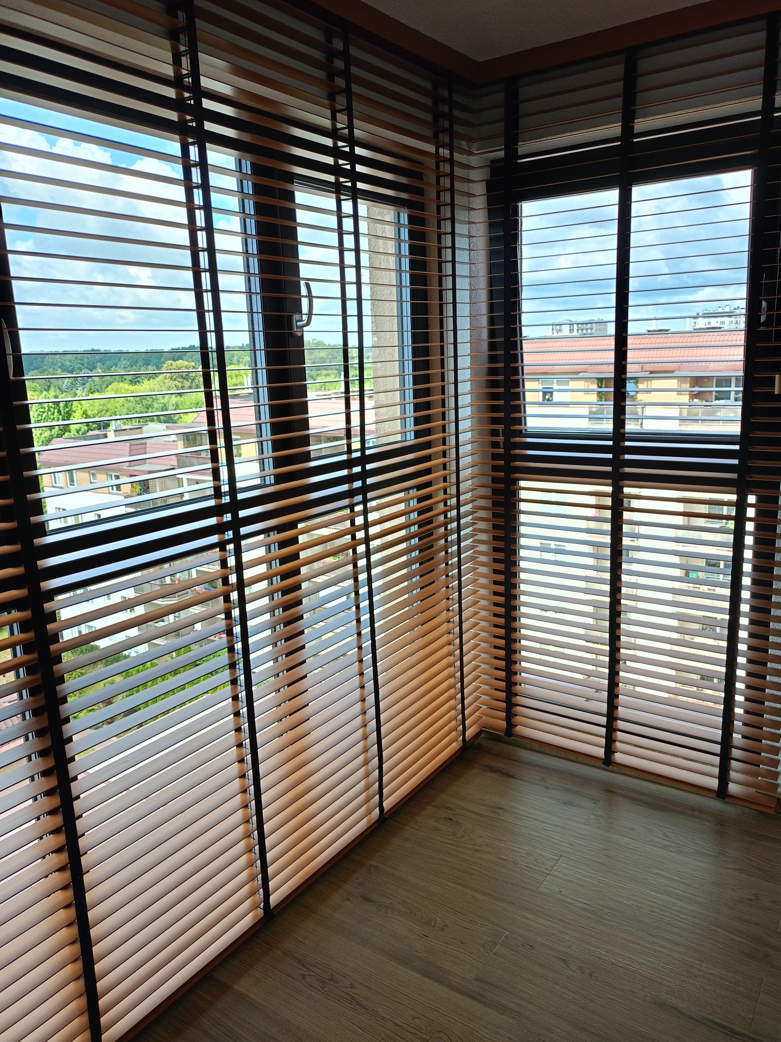Beżowe żaluzje drewniane z ciemnymi ramami okiennymi, widok na zieleń i budynki przez okno, parkiet na podłodze, wnętrze z naturalnym światłem.