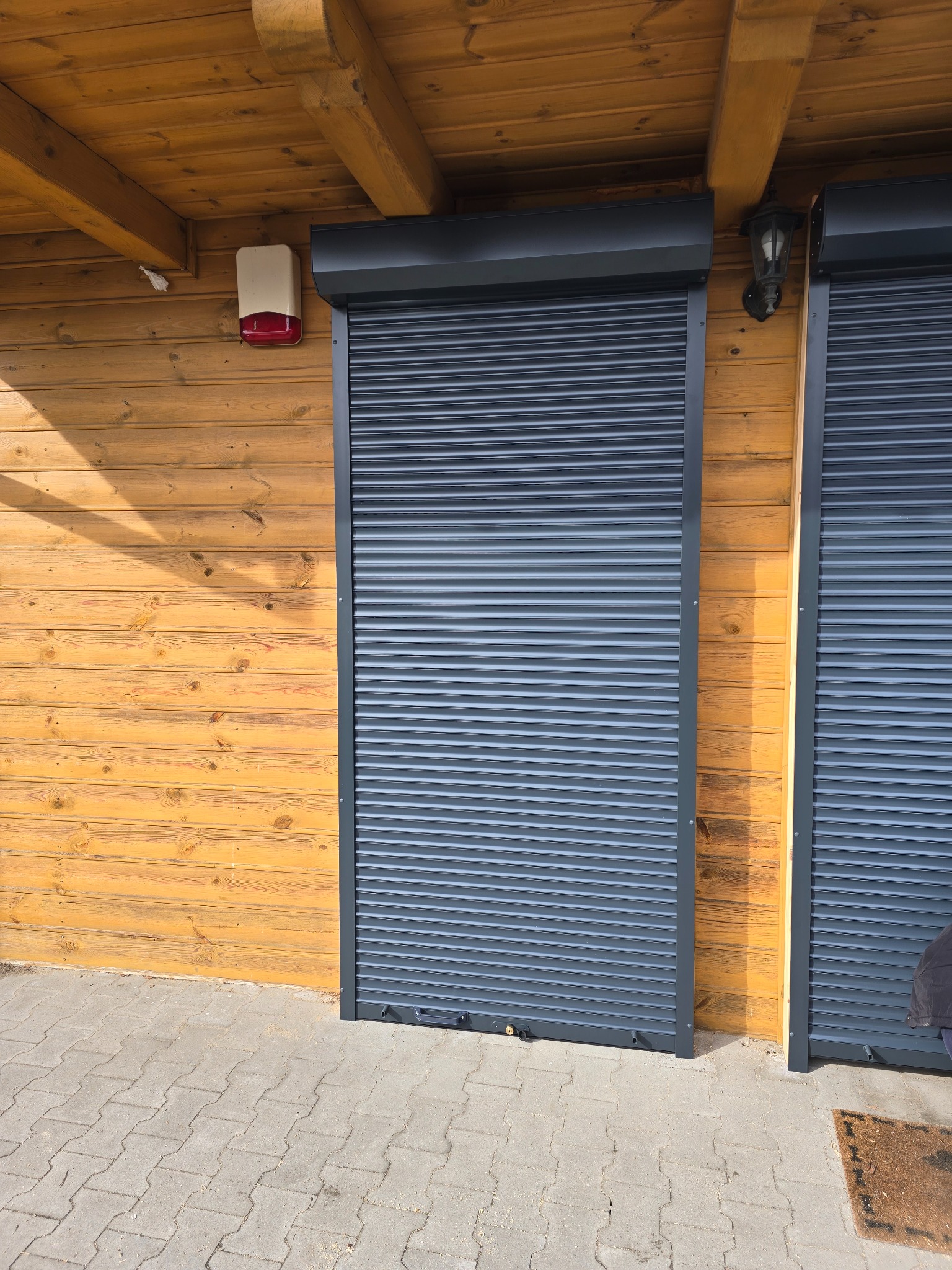 Zamknięta, grafitowa roleta zewnętrzna na drewnianej elewacji budynku z alarmem i lampą. Widoczna kostka brukowa i fragment drugiej rolety.