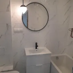 Nowoczesna łazienka z białymi marmurkowymi płytkami, okrągłym lustrem w czarnej ramie, podwieszaną szafką z umywalką i czarną baterią oraz wiszącą lampą z kulistym kloszem.