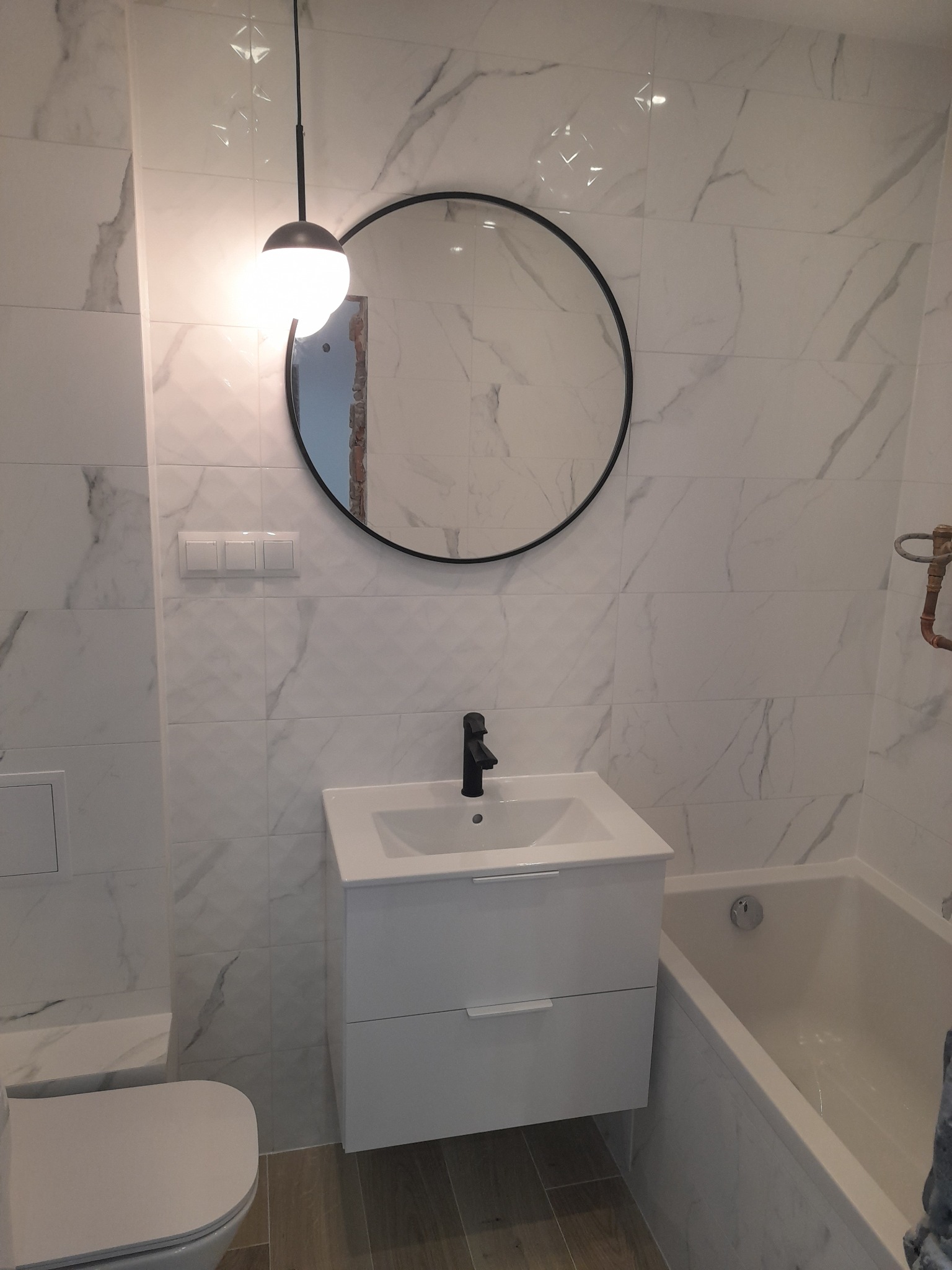 Nowoczesna łazienka z białymi marmurkowymi płytkami, okrągłym lustrem w czarnej ramie, podwieszaną szafką z umywalką i czarną baterią oraz wiszącą lampą z kulistym kloszem.