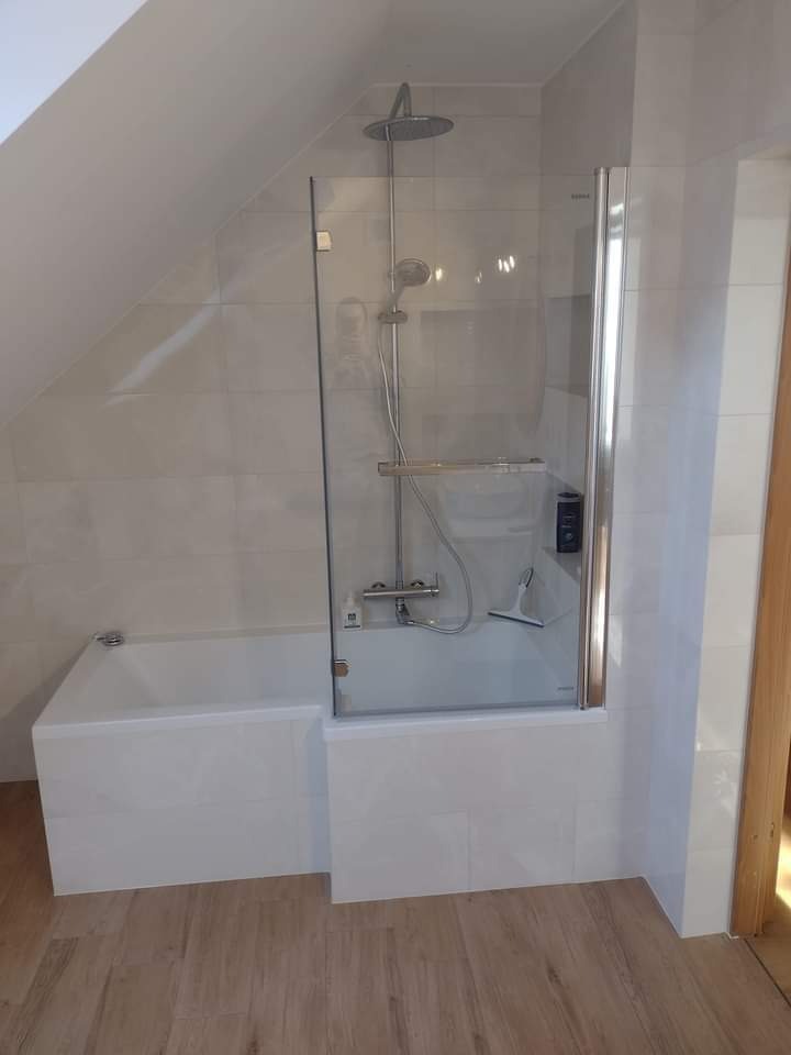 Biała wanna z przeszklonym parawanem nawannowym w łazience na poddaszu, wykończonej płytkami w odcieniach bieli i beżu.