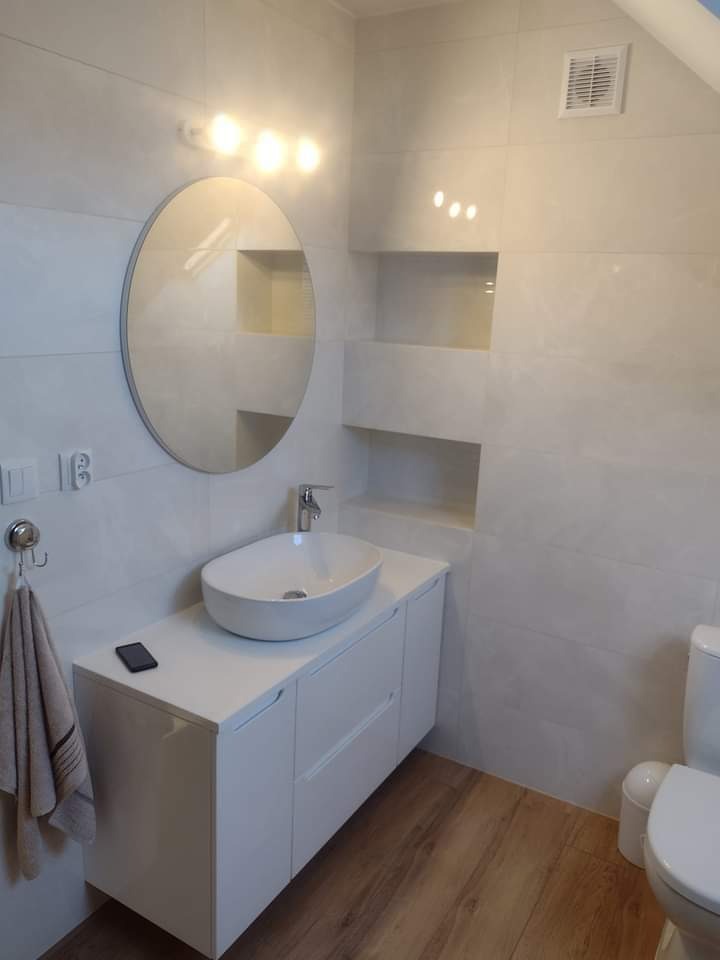 Nowoczesna łazienka z owalną umywalką nablatową na białej szafce, okrągłym lustrem podświetlanym trzema żarówkami i płytkami imitującymi marmur z wnękami.