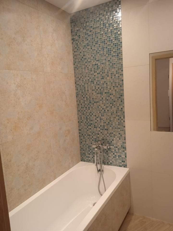 Biała wanna obudowana beżowymi płytkami, ściana z mozaiki w odcieniach turkusu i beżu, beżowe płytki na ścianach i podłodze, bateria wannowa dwuuchwytowa.