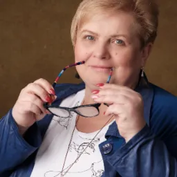 Portret uśmiechniętej kobiety w niebieskiej marynarce, trzymającej okulary z kolorowymi zausznikami, z widocznym naszyjnikiem z dużym kamieniem i wzorem baletnic na koszulce.