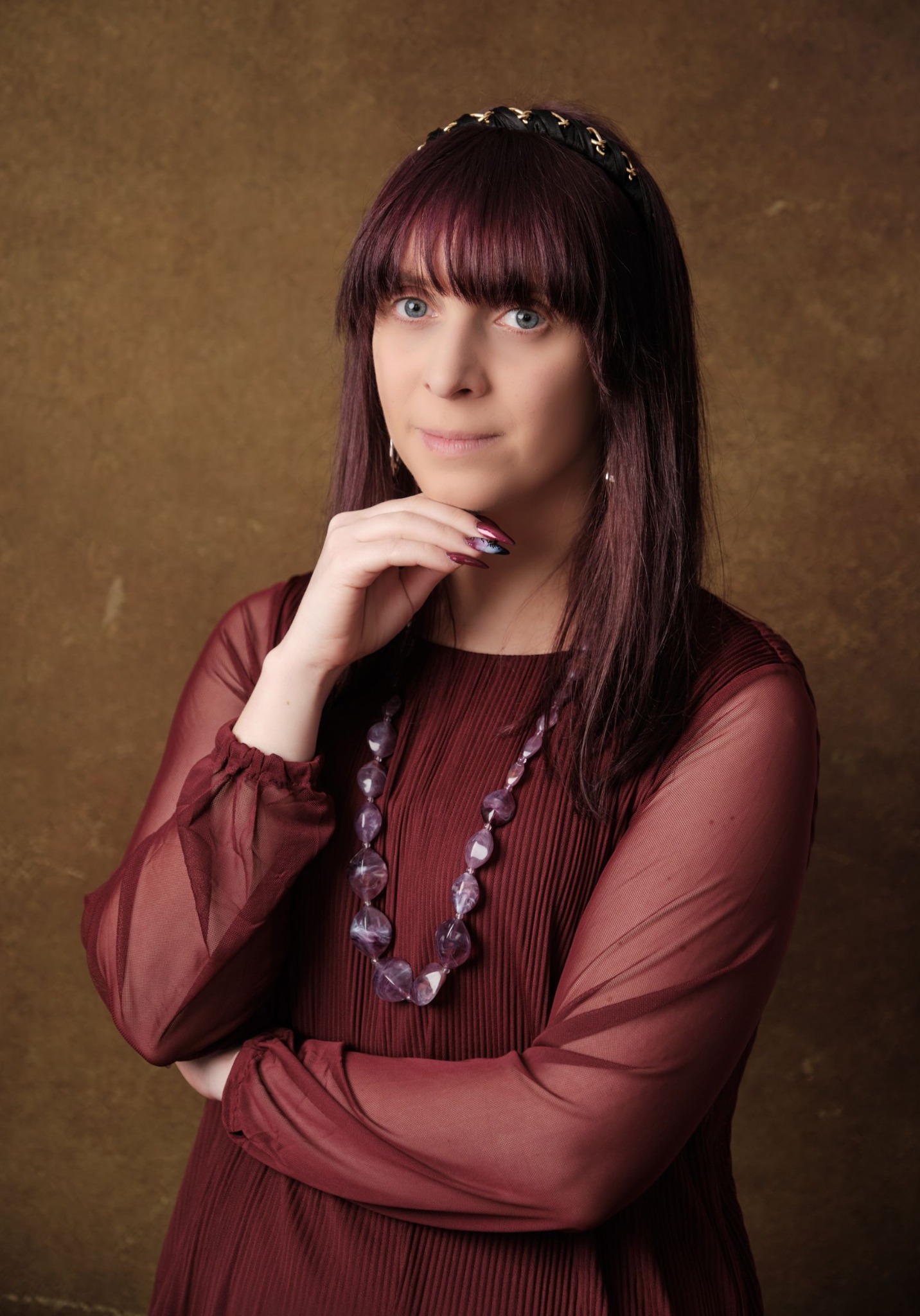 Portret kobiety z fioletowymi włosami, w bordowej sukience i z naszyjnikiem z dużych, fioletowych kamieni, pozującej na rozmytym, złotym tle.