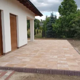 Nowoczesny taras z jasnej kostki brukowej ułożonej przy białym domu z brązowymi oknami i roletami, obrzeża wykończone ciemniejszą kostką, widok na ogród.