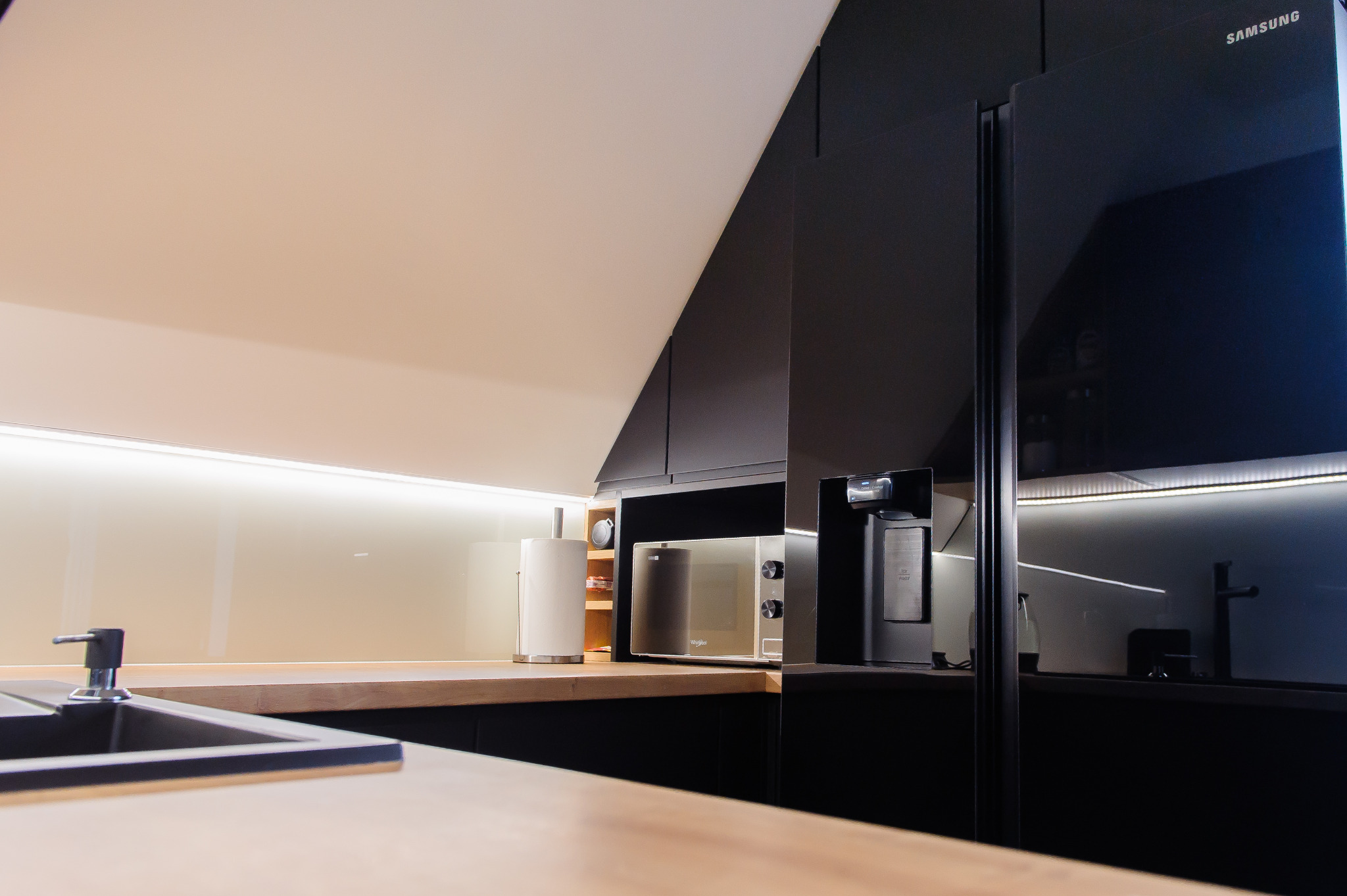 Nowoczesna kuchnia z czarnymi szafkami, drewnianym blatem, białym panelem ściennym z podświetleniem LED, wbudowaną mikrofalówką Whirlpool i lodówką Samsung z dystrybutorem wody.