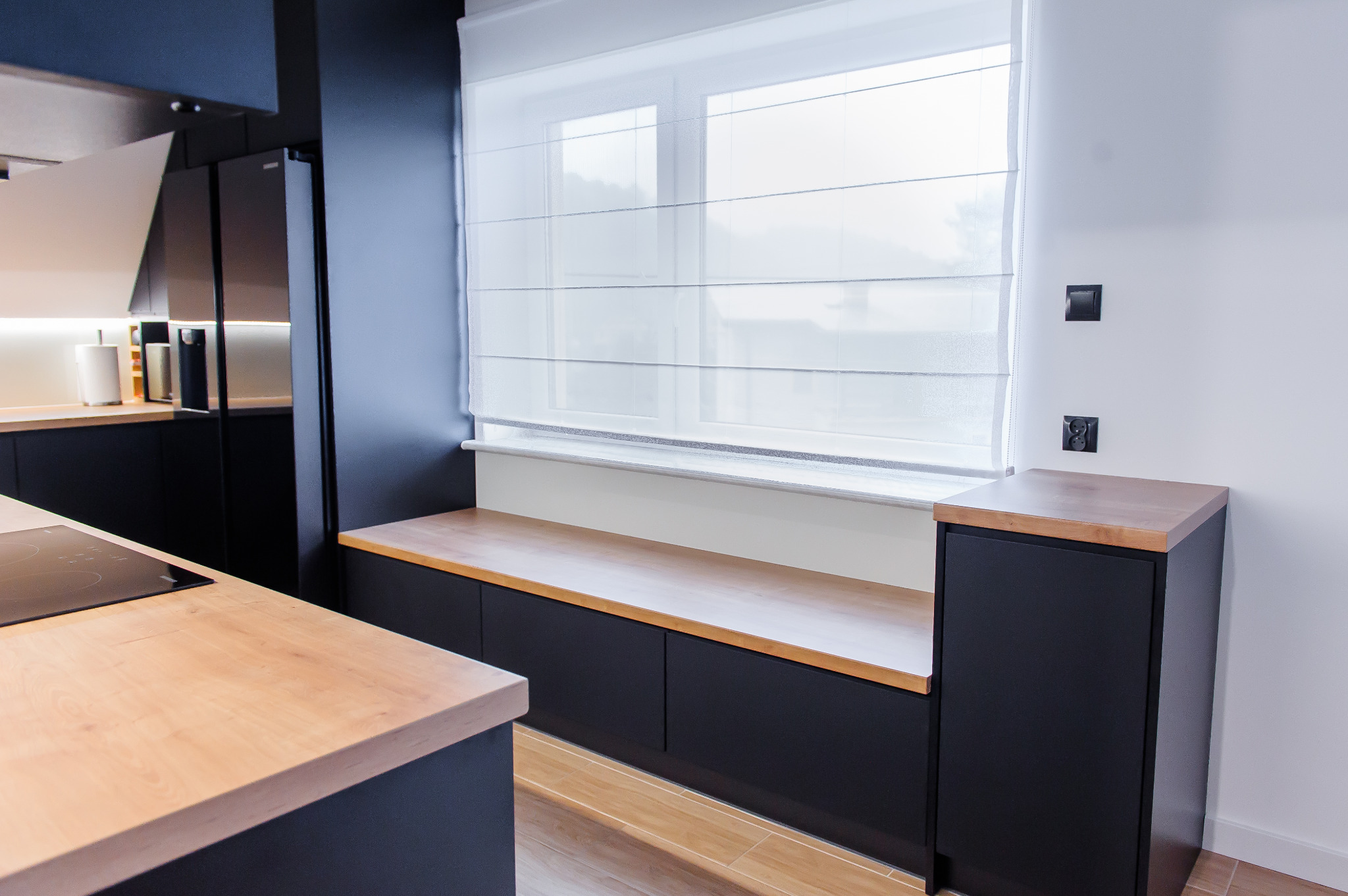 Nowoczesna kuchnia z czarnymi szafkami i drewnianym blatem, widok na okno z roletami rzymskimi, jasne drewno na podłodze i blacie, minimalistyczne gniazdka elektryczne na białej ścianie.