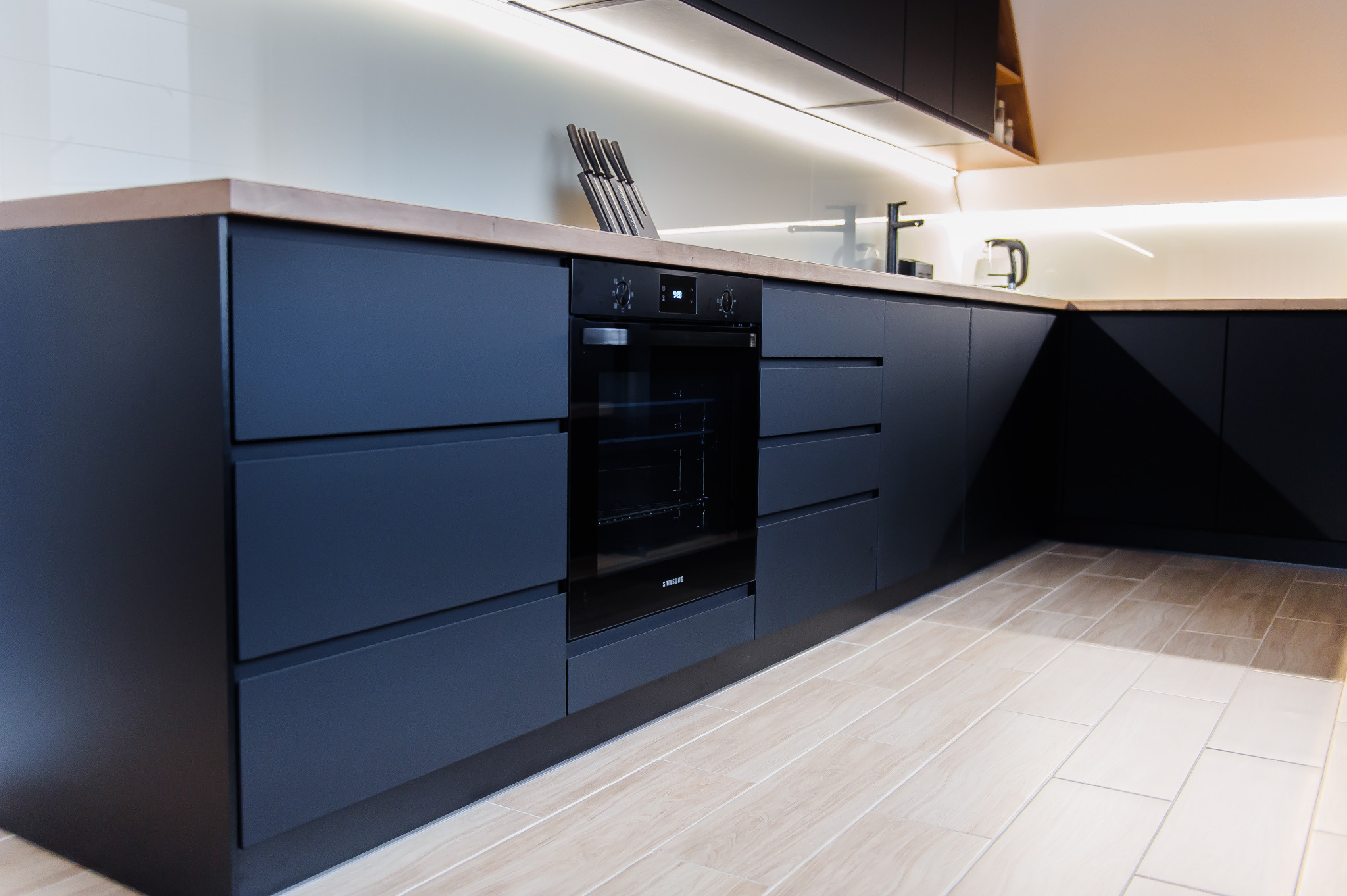 Nowoczesna kuchnia z matowymi, grafitowymi frontami szafek, drewnianym blatem i wbudowanym piekarnikiem Samsung, oświetlona listwą LED pod szafkami wiszącymi.