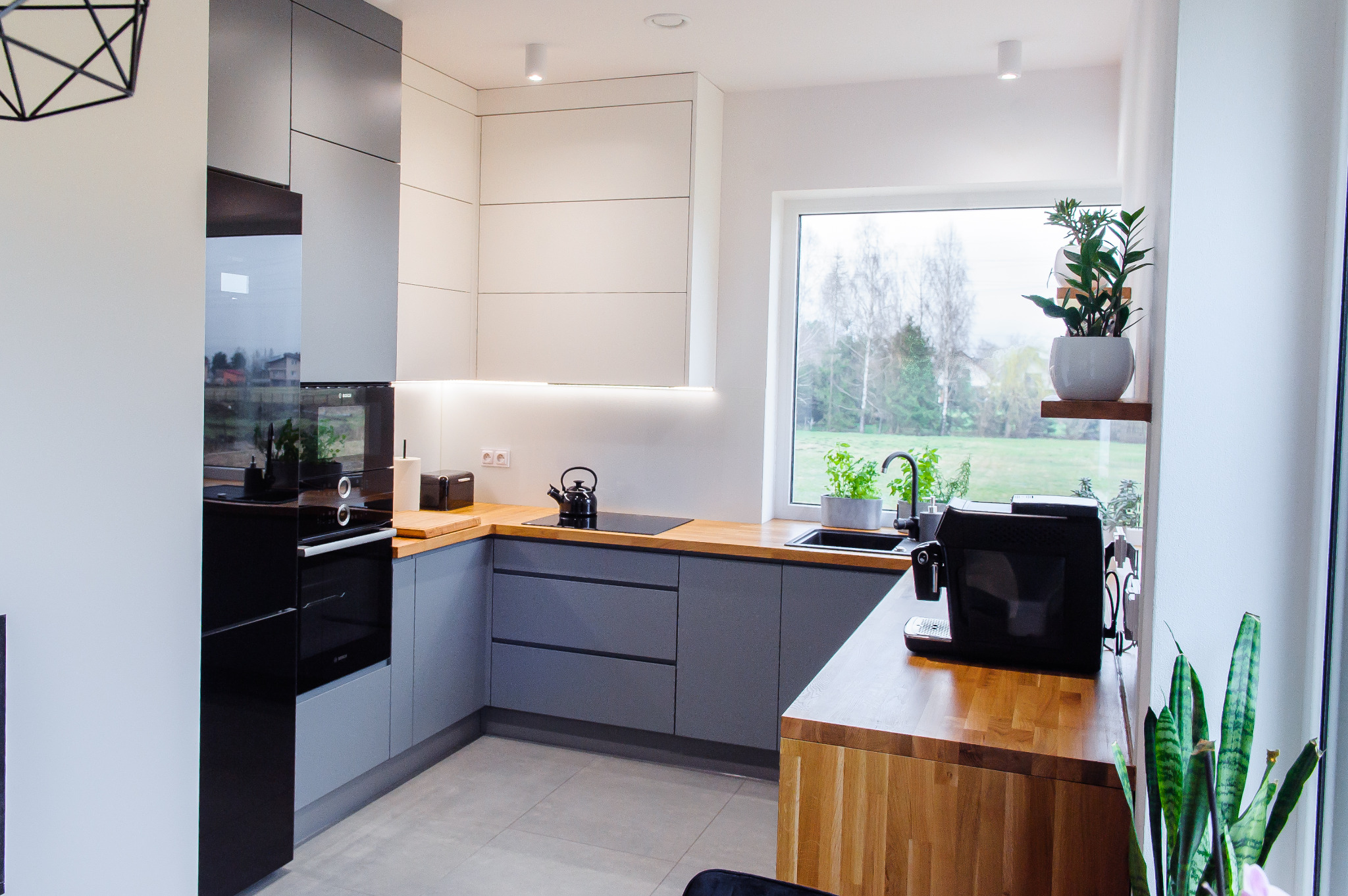 Nowoczesna kuchnia w szaro-białej tonacji z drewnianymi blatami, wyposażona w czarny piekarnik, płytę indukcyjną i ekspres do kawy. Widok na ogród przez okno.