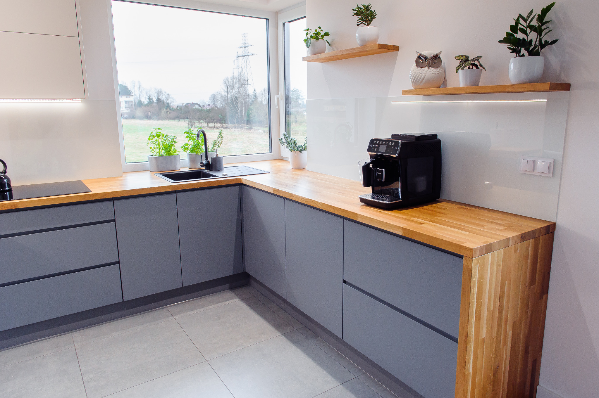 Szara kuchnia na wymiar z drewnianym blatem, czarnym zlewem i ekspres do kawy na blacie, widok na zielone pole za oknem.