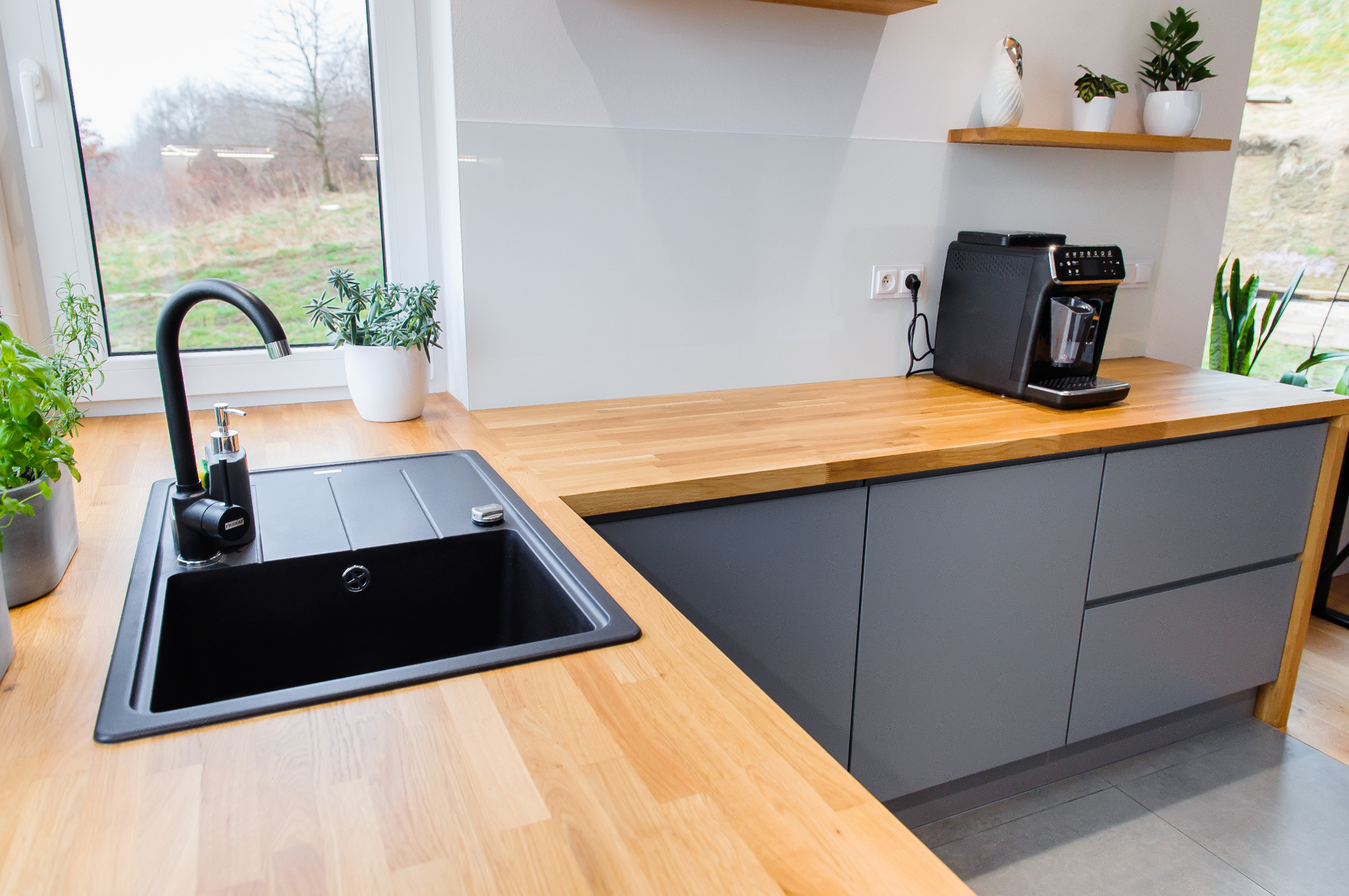 Nowoczesna kuchnia z drewnianym blatem, szarymi szafkami i czarnym zlewem Franke z dozownikiem, oświetlona naturalnym światłem z okna z widokiem na zieleń. Na blacie ekspres do kawy i doniczki...