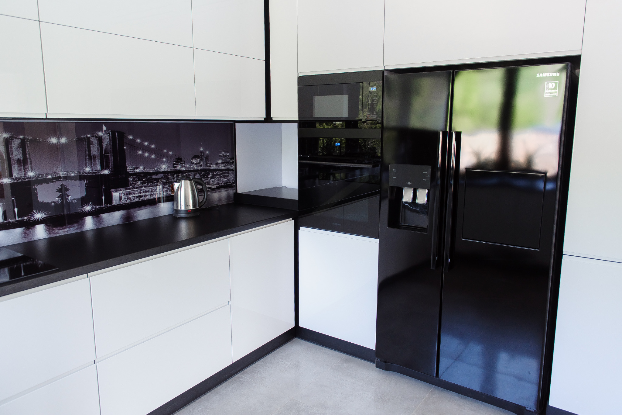 Nowoczesna kuchnia z białymi frontami i czarnym blatem, na którym stoi czajnik elektryczny. Na ścianie fototapeta z nocnym widokiem miasta. W zabudowie czarna lodówka side-by-side i piekarnik.