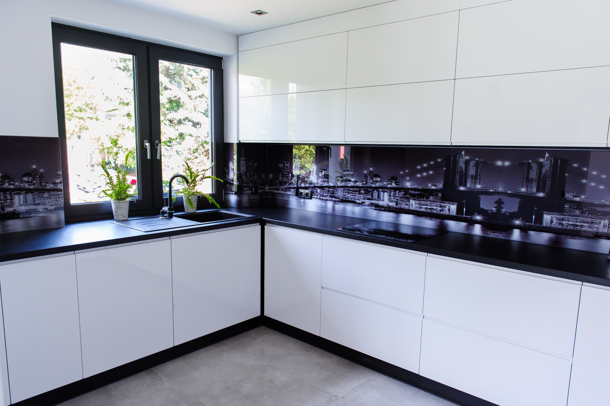 Biała, nowoczesna kuchnia z czarnym blatem i fototapetą z motywem nocnego miasta nad blatem, rośliny w doniczkach przy oknie.