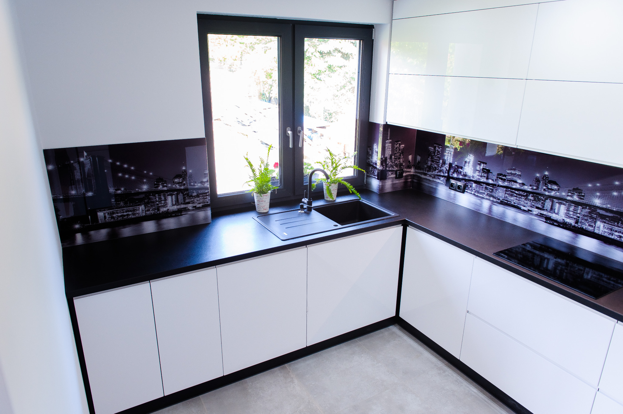 Nowoczesna kuchnia z białymi, lakierowanymi frontami, czarnym blatem i czarnym zlewozmywakiem, ozdobiona grafiką z motywem nocnego miasta na ścianie między szafkami, z dwoma paprotkami w doniczkach...