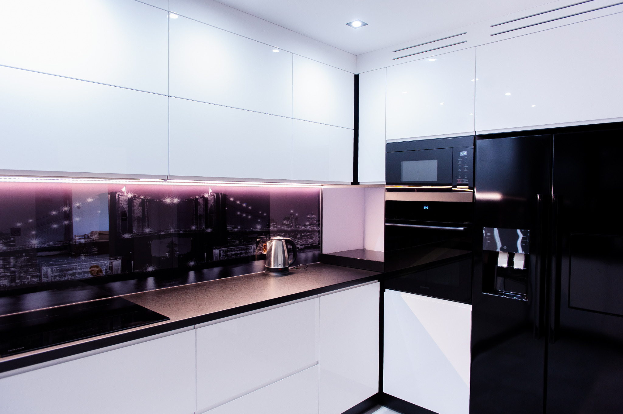 Nowoczesna kuchnia z białymi, gładkimi frontami szafek, czarnym blatem i panelem nad blatem z motywem panoramy miasta podświetlonym fioletowym światłem LED, obok czarna lodówka side-by-side.