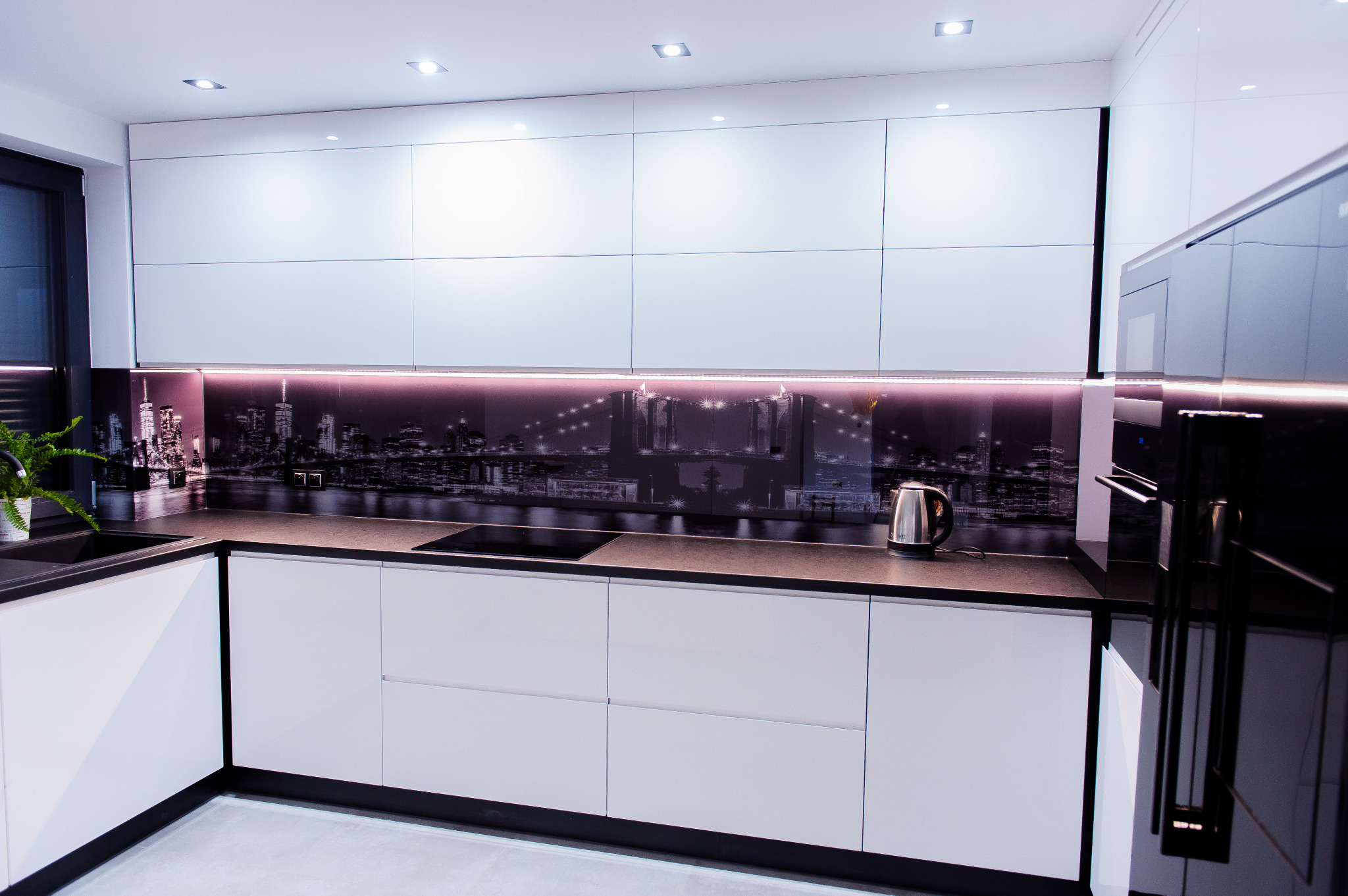 Nowoczesna kuchnia z białymi, gładkimi frontami szafek, czarnym blatem i panelem szklanym z motywem panoramy miasta podświetlonym od dołu, widoczne wbudowane sprzęty AGD i czajnik.