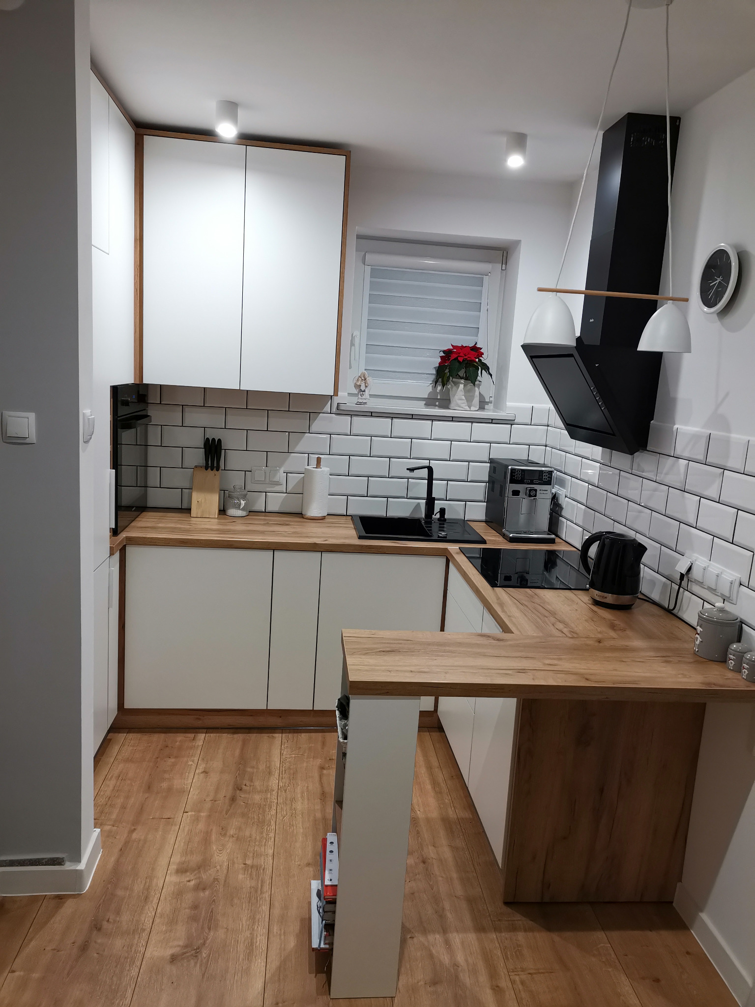 Nowoczesna, narożna kuchnia z białymi szafkami i drewnianym blatem, czarną armaturą i okapem, wykończona białymi płytkami w stylu metro, oświetlona wiszącymi lampami.