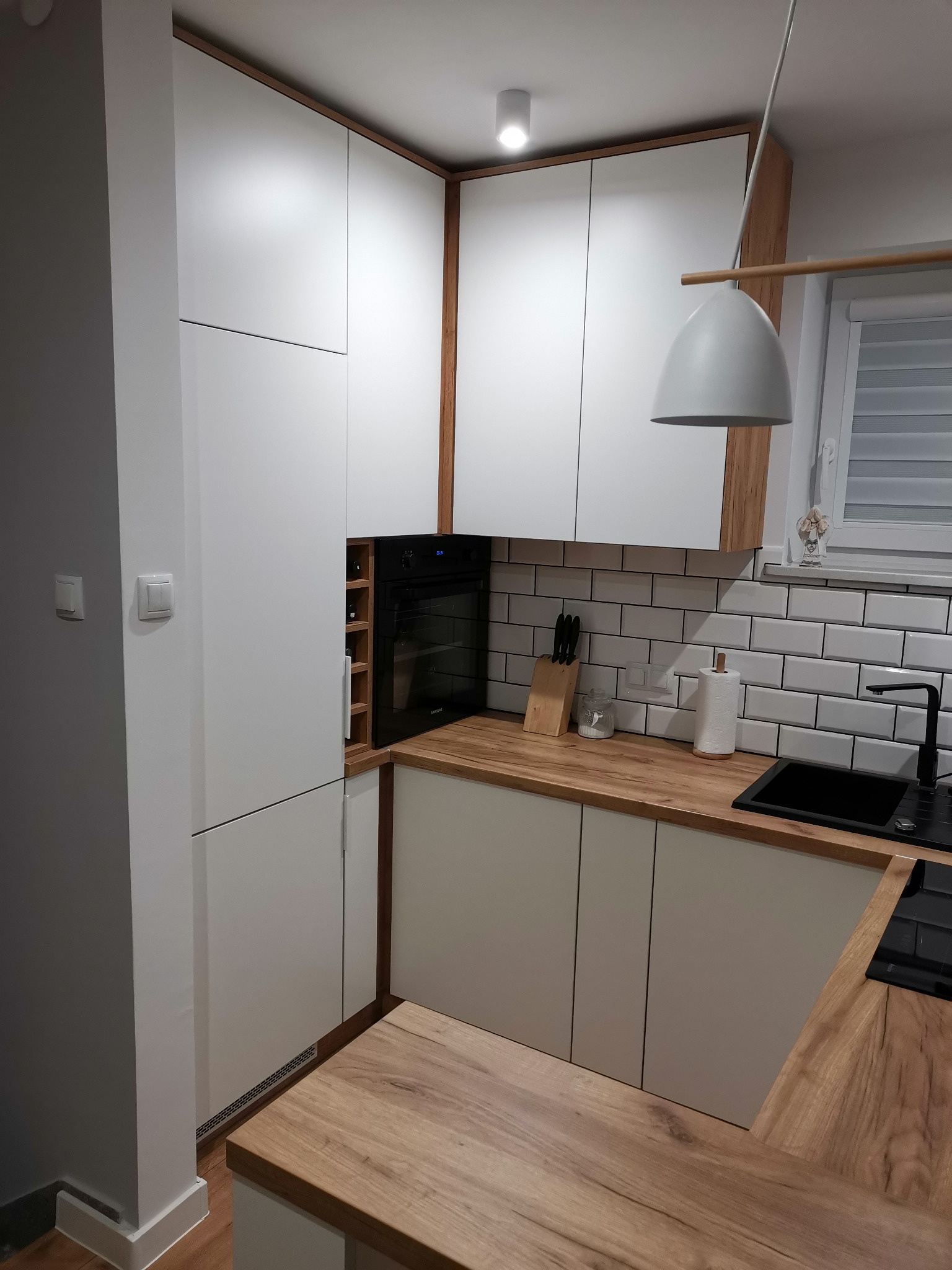 Nowoczesna kuchnia z białymi szafkami, drewnianymi akcentami, czarnym zlewem i piekarnikiem w zabudowie. Widoczny drewniany blat, białe płytki na ścianie i minimalistyczna lampa wisząca.