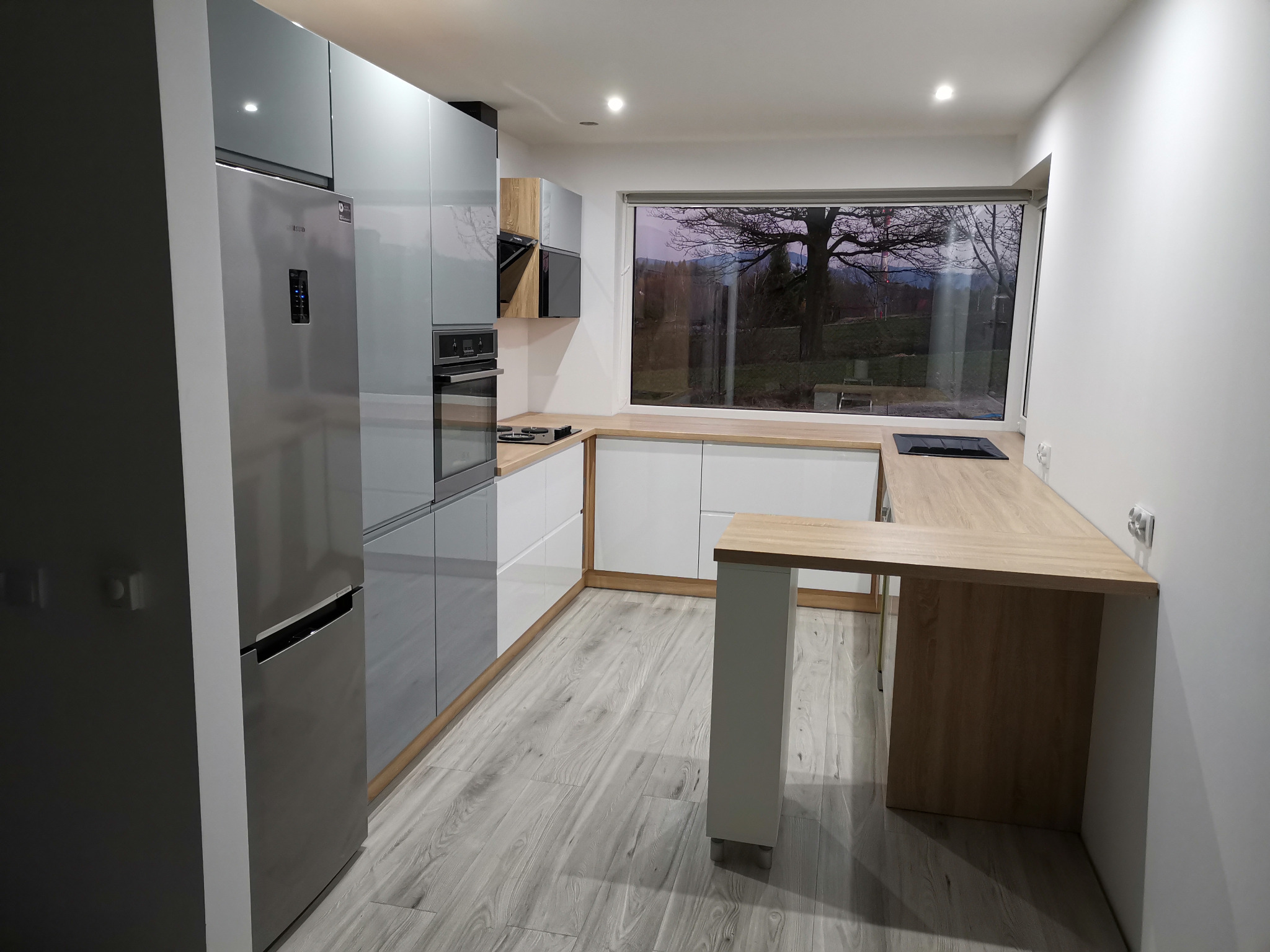 Nowoczesna kuchnia z szarymi i białymi szafkami, drewnianym blatem i dużym oknem z widokiem na drzewo.