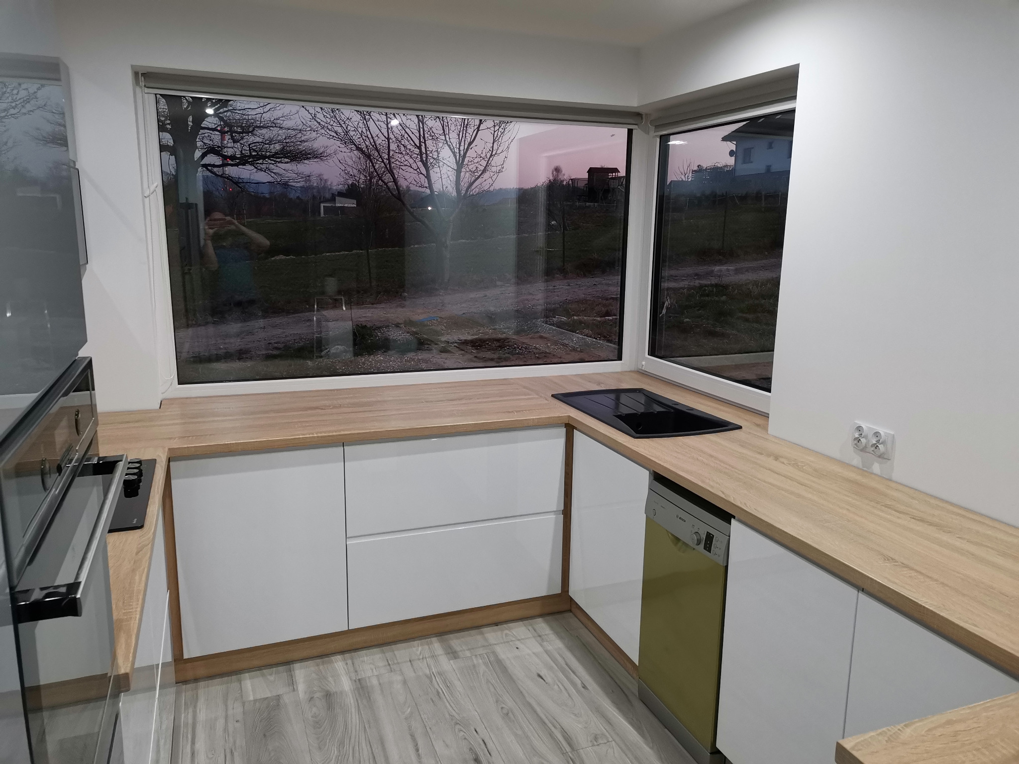 Nowoczesna kuchnia z białymi frontami szafek i blatem w kolorze jasnego drewna, z widokiem na ogród przez duże okno, z wbudowaną płytą grzewczą i piekarnikiem.