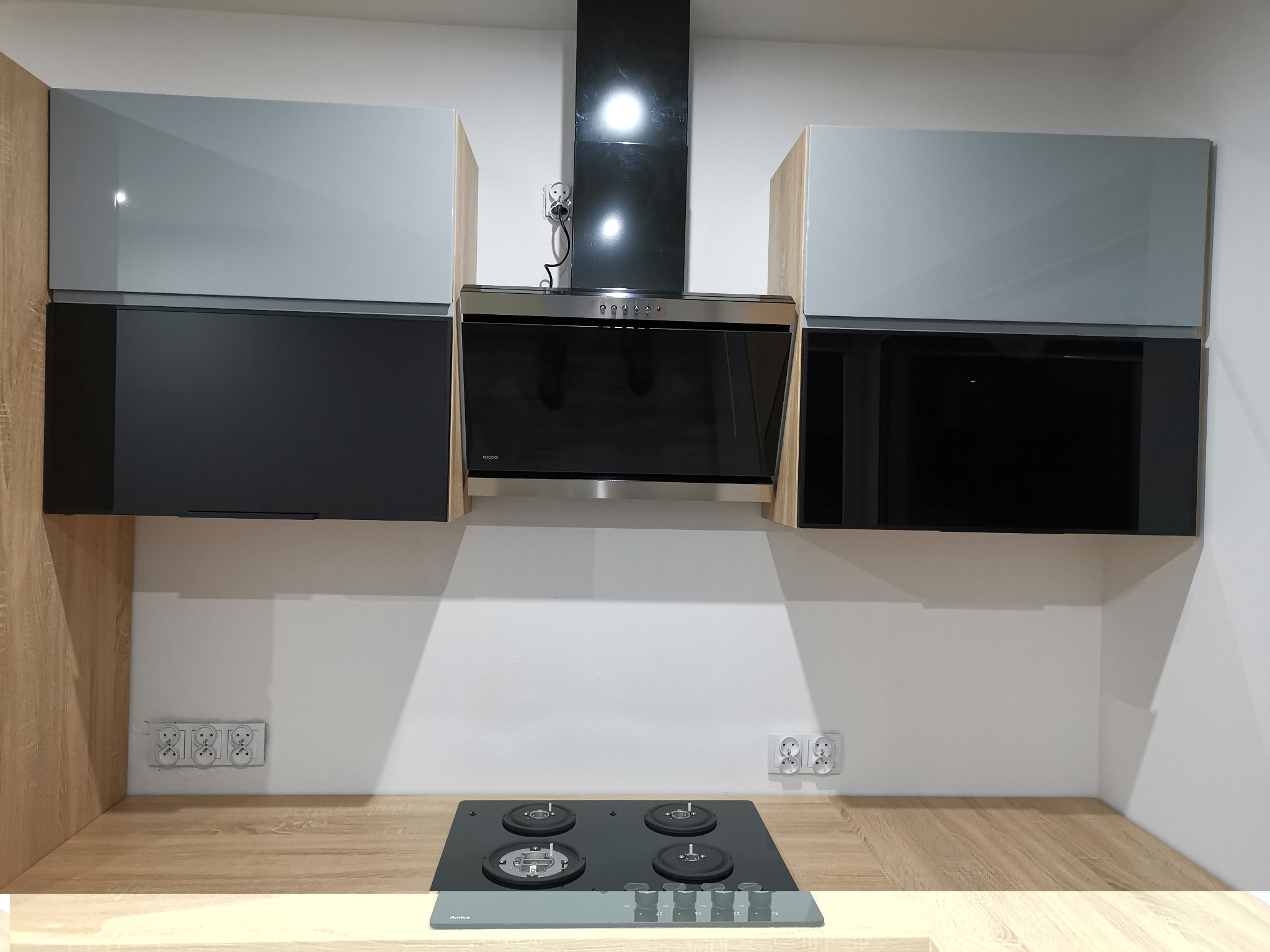 Nowoczesna kuchnia: górne szafki z szarymi i czarnymi frontami, drewnopodobny blat, czarna płyta gazowa i okap.