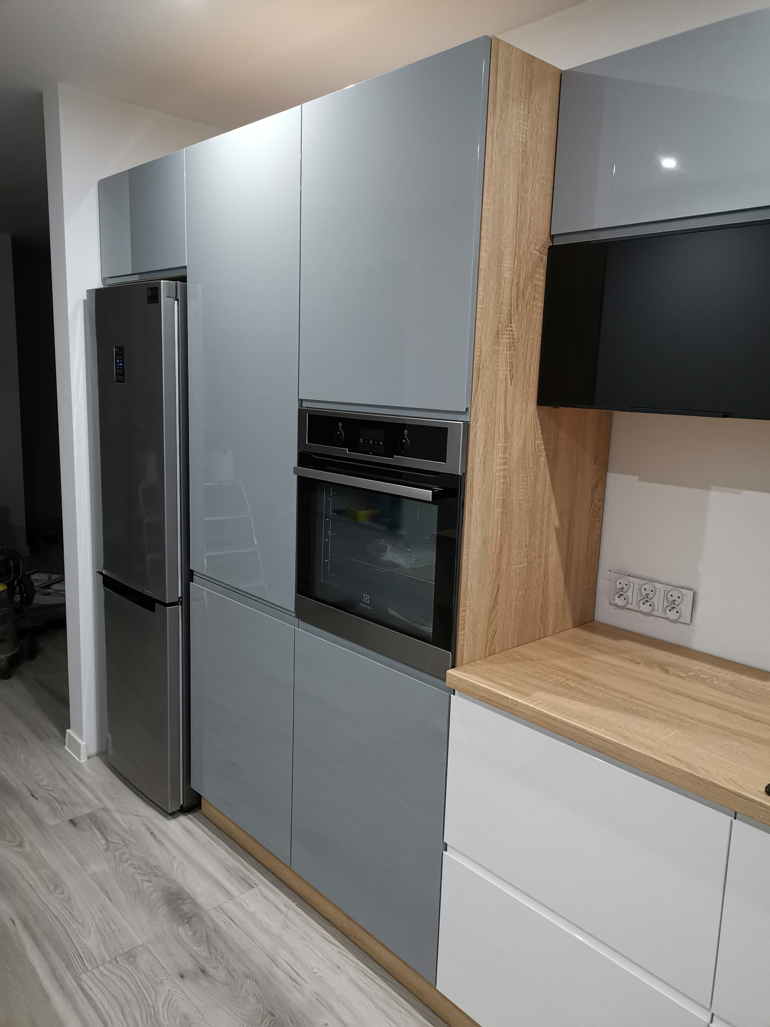 Nowoczesna kuchnia z szarymi, lakierowanymi frontami, drewnianymi akcentami i srebrną lodówką side-by-side, z piekarnikiem w zabudowie i jasnym blatem.