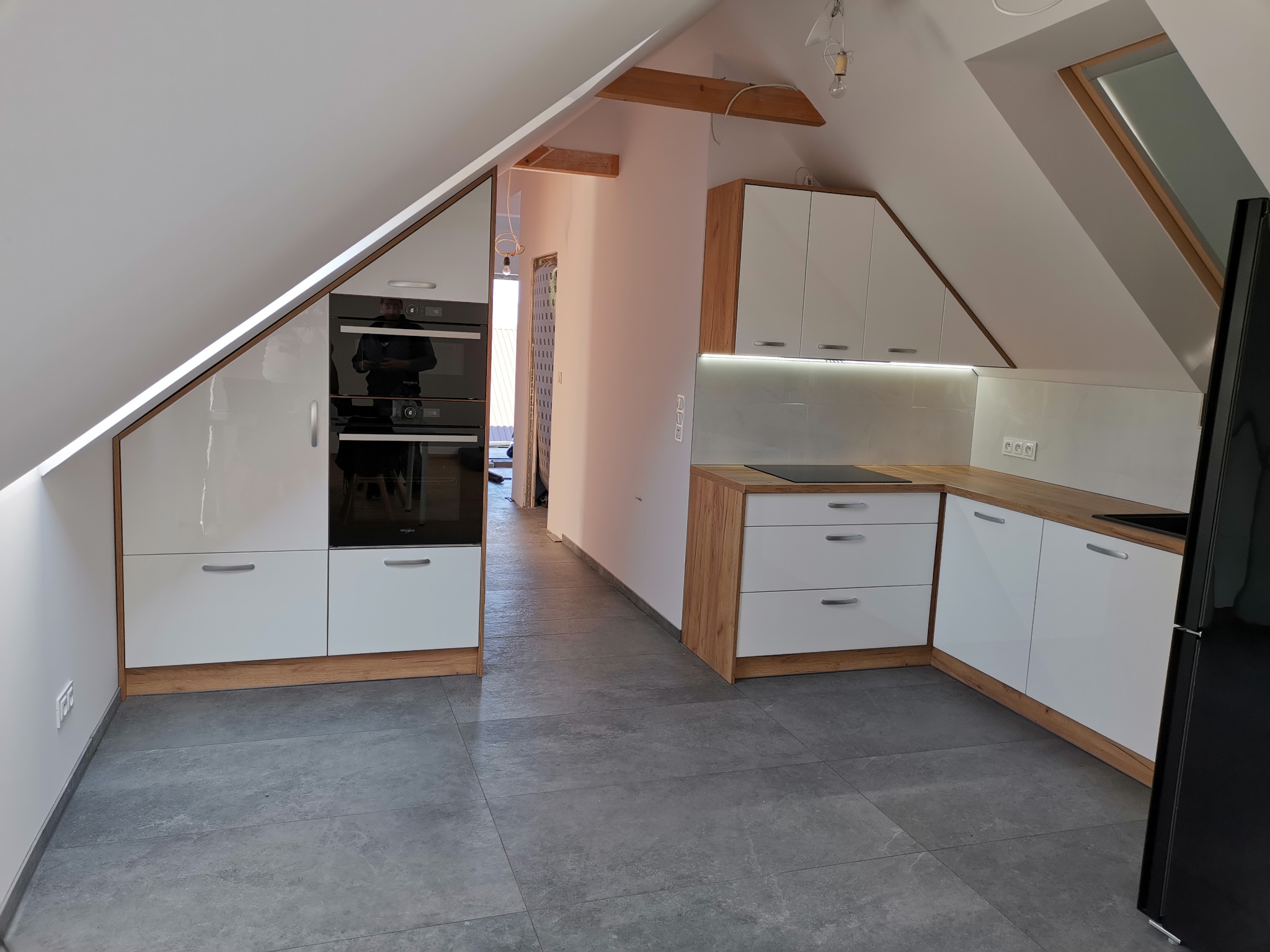 Nowoczesna kuchnia w skosach poddasza z białymi frontami i drewnianymi akcentami, widoczny sprzęt AGD w zabudowie i szara podłoga.