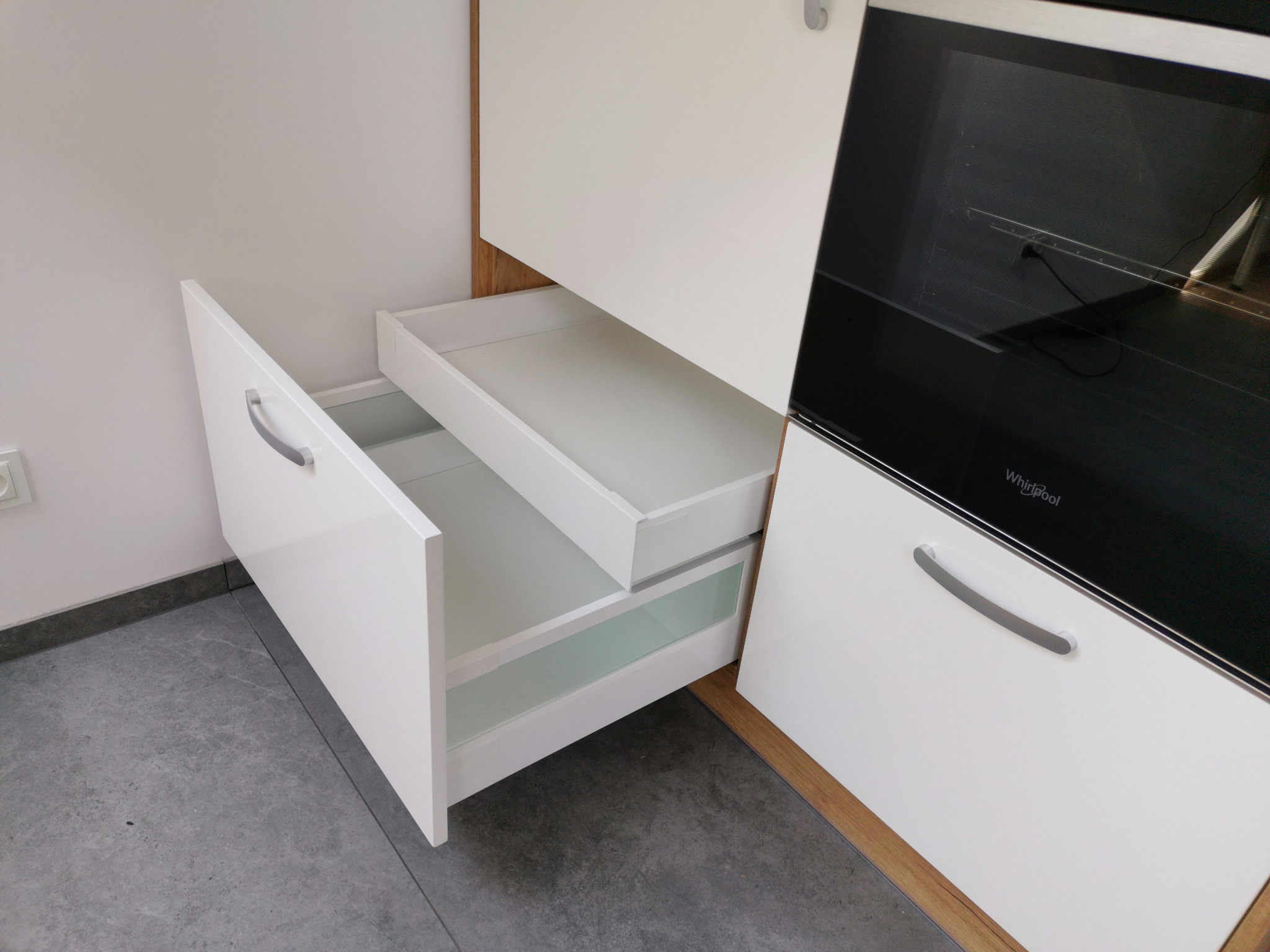 Biała kuchnia na wymiar z otwartymi szufladami, widoczny front szuflady z uchwytem i przeszklonym bokiem, obok zabudowana czarna piekarnik Whirlpool.
