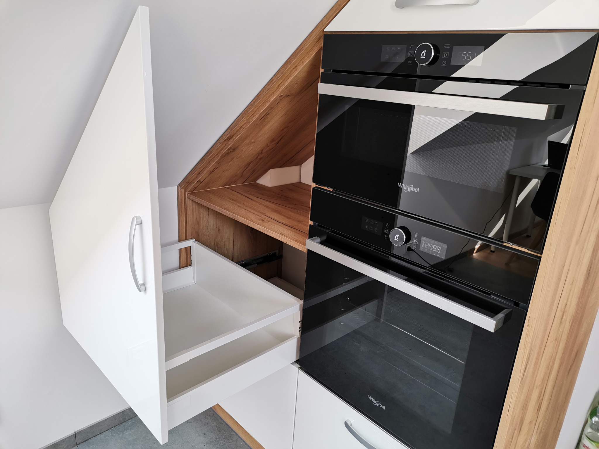 Biała zabudowa kuchenna pod skosem z otwartą szufladą i wbudowanym piekarnikiem oraz mikrofalówką marki Whirlpool, wykończona drewnopodobnym blatem.