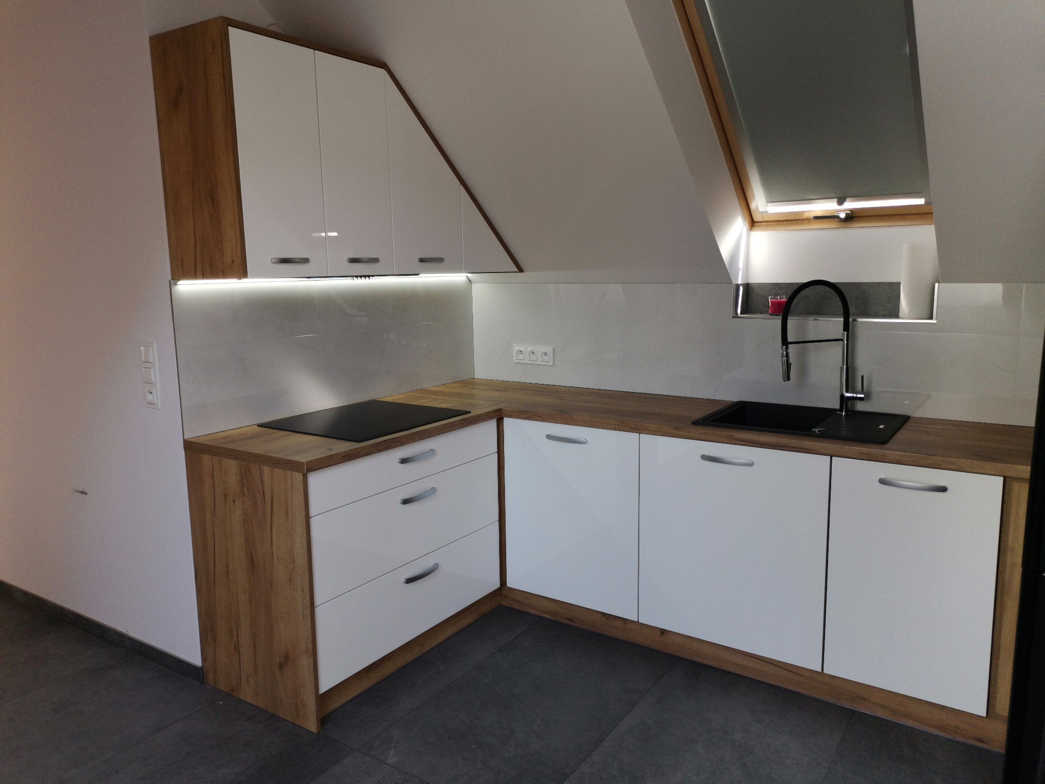 Nowoczesna kuchnia w skosach: białe fronty szafek dolnych i górnych z drewnianymi korpusami, blat imitujący drewno, czarny zlew i bateria, płyta indukcyjna, okno dachowe.