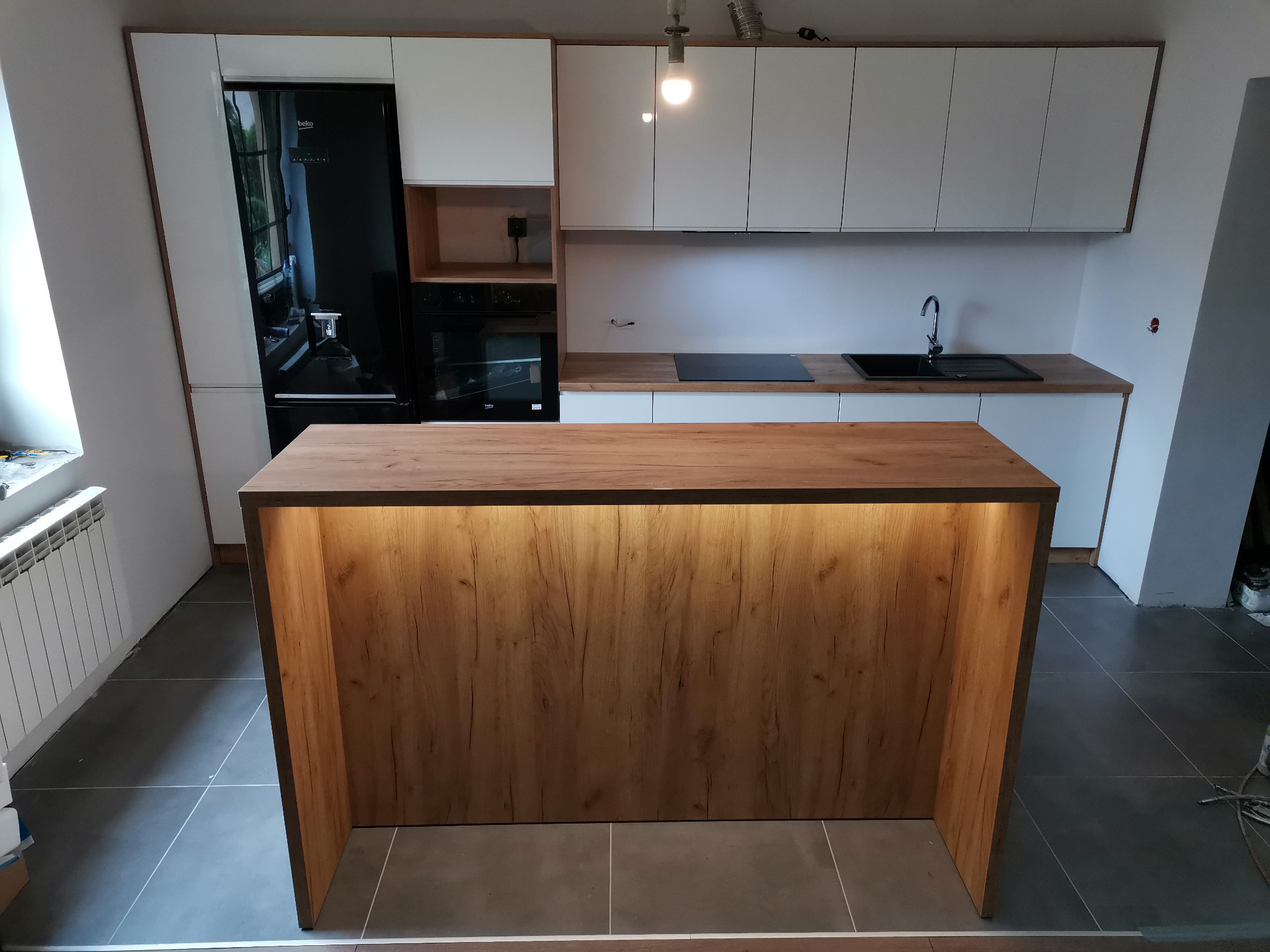 Nowoczesna kuchnia z białymi szafkami i drewnianym blatem wyspowym z podświetleniem, czarny piekarnik i lodówka w zabudowie, szare płytki podłogowe.