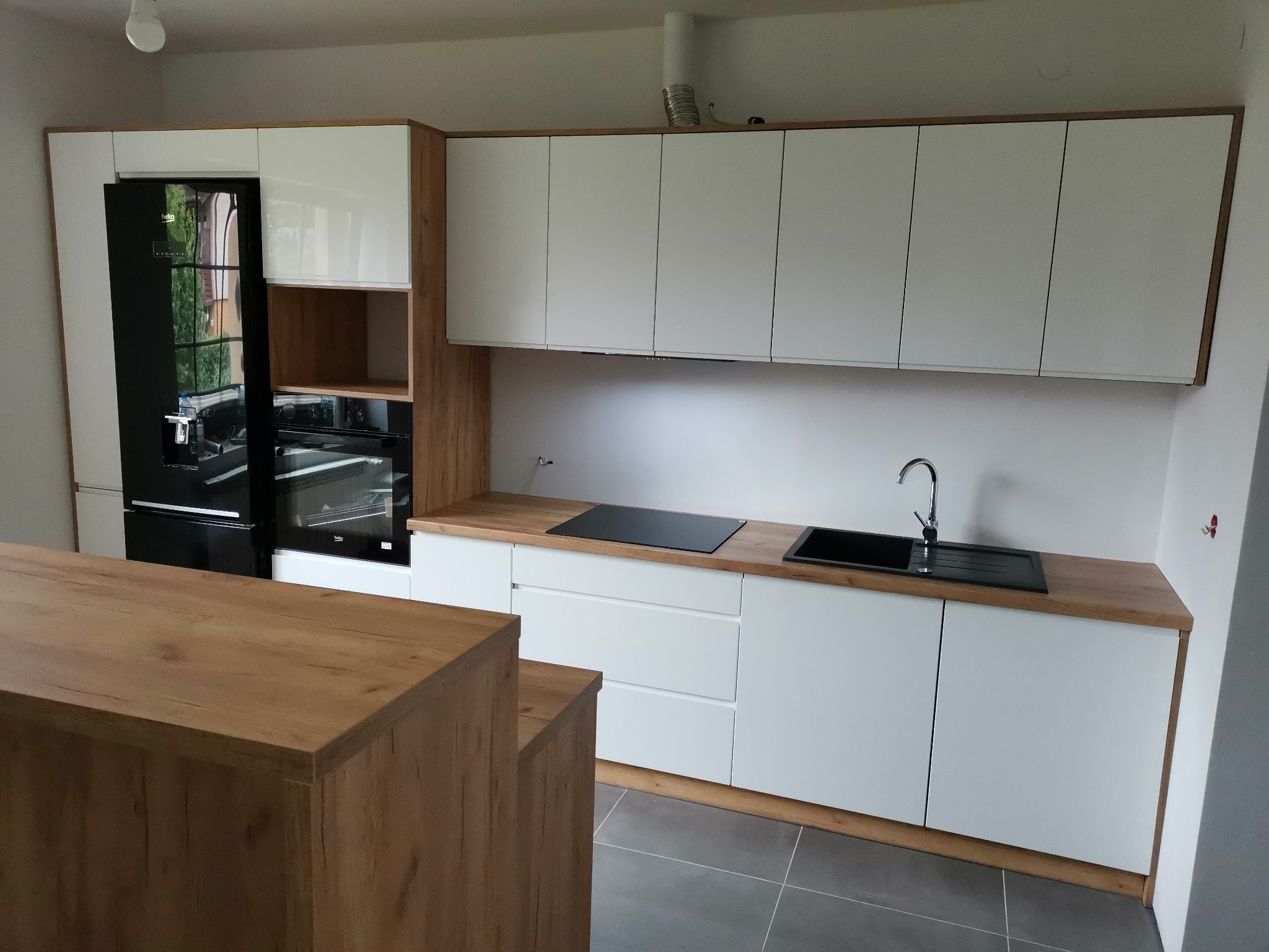 Nowoczesna kuchnia z białymi frontami szafek, drewnianymi blatami i wykończeniami, czarnym zlewozmywakiem i płytą indukcyjną, oraz dwupoziomową wyspą kuchenną z drewnianym blatem.