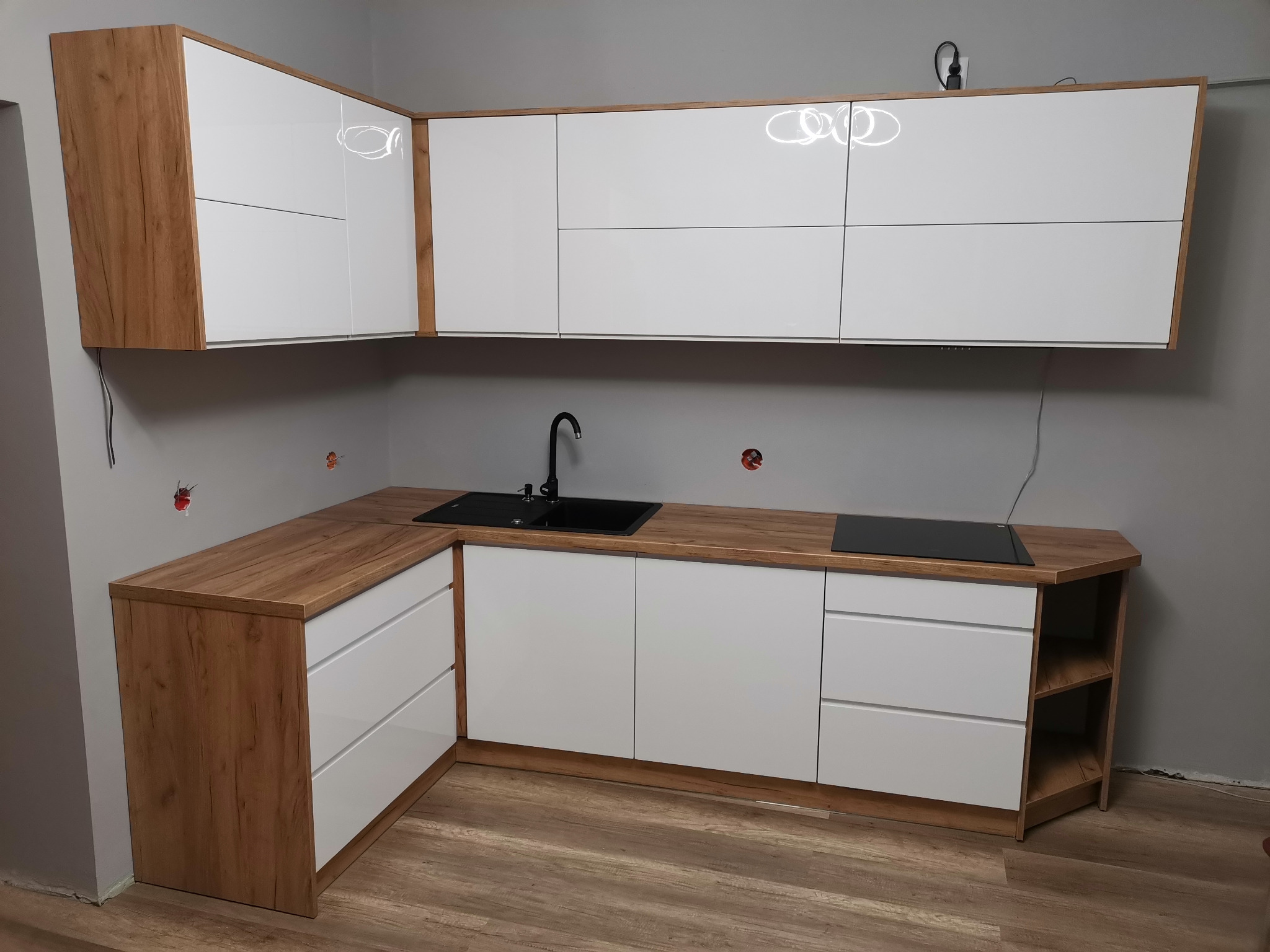 Nowoczesna kuchnia narożna z białymi, błyszczącymi frontami i blatem imitującym drewno, czarny zlew i płyta indukcyjna, szare ściany w trakcie wykończenia.