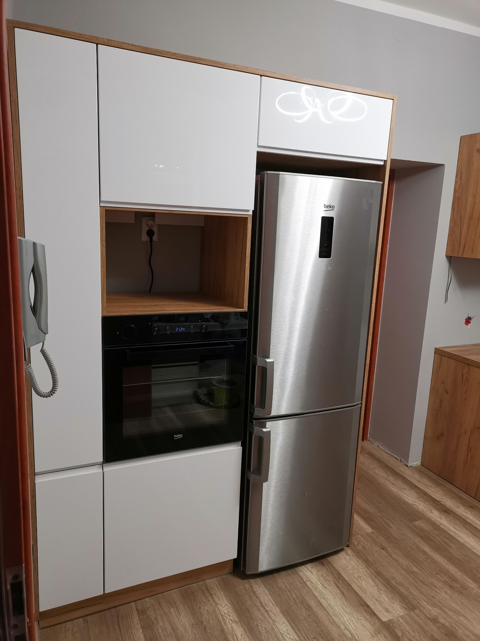 Białe szafki kuchenne z drewnianymi akcentami, wbudowany piekarnik Beko, srebrna lodówka Beko, szary interkom na ścianie, drewnopodobna podłoga.