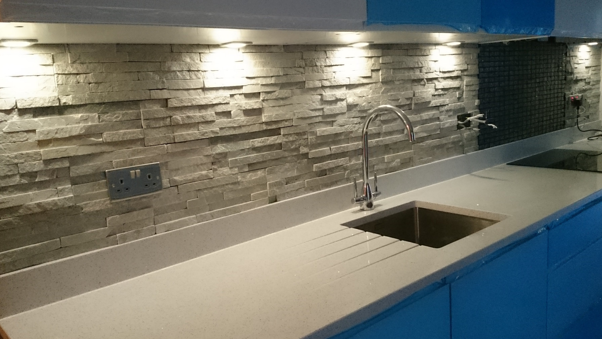 Wykończona ściana nad blatem kuchennym z dekoracyjną okładziną imitującą kamień, podświetlana punktowymi lampami LED, z widocznym chromowanym kranem i gniazdkiem elektrycznym.