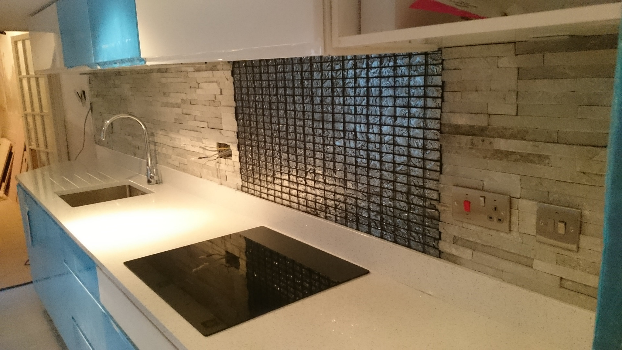 Fragment kuchni z białym blatem, indukcyjną płytą grzewczą i zlewozmywakiem, ściana wyłożona częściowo mozaiką i kamieniem dekoracyjnym, widoczne gniazdka elektryczne.