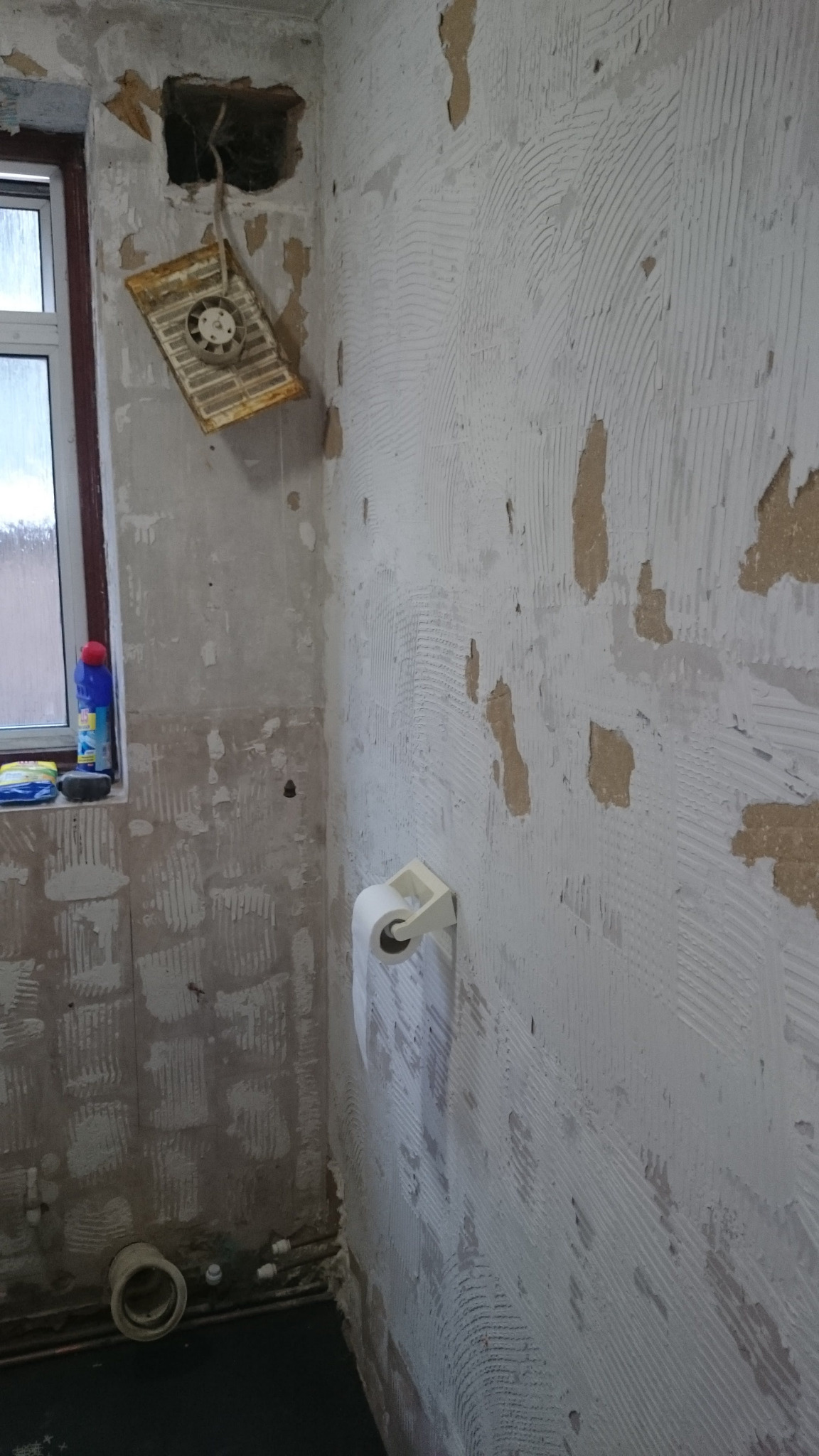 Łazienka w trakcie przygotowania do remontu: ściany z resztkami starej farby i kleju, widoczny otwór po wentylacji z wiszącą kratką, okno z butelką płynu do czyszczenia i rolka papieru toaletowego.