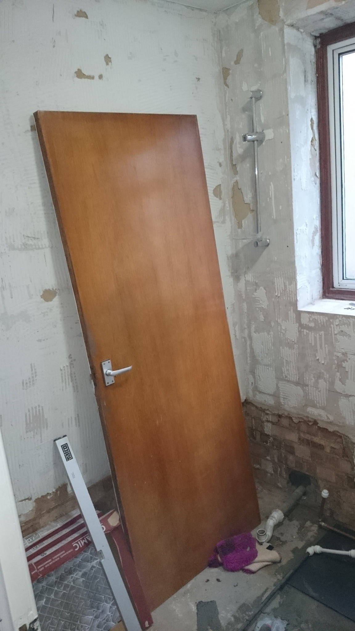 Demontaż starej drewnianej futryny w łazience podczas prac remontowych, widoczne ślady po płytkach na ścianach i rurach, karton z płytkami pod ścianą.