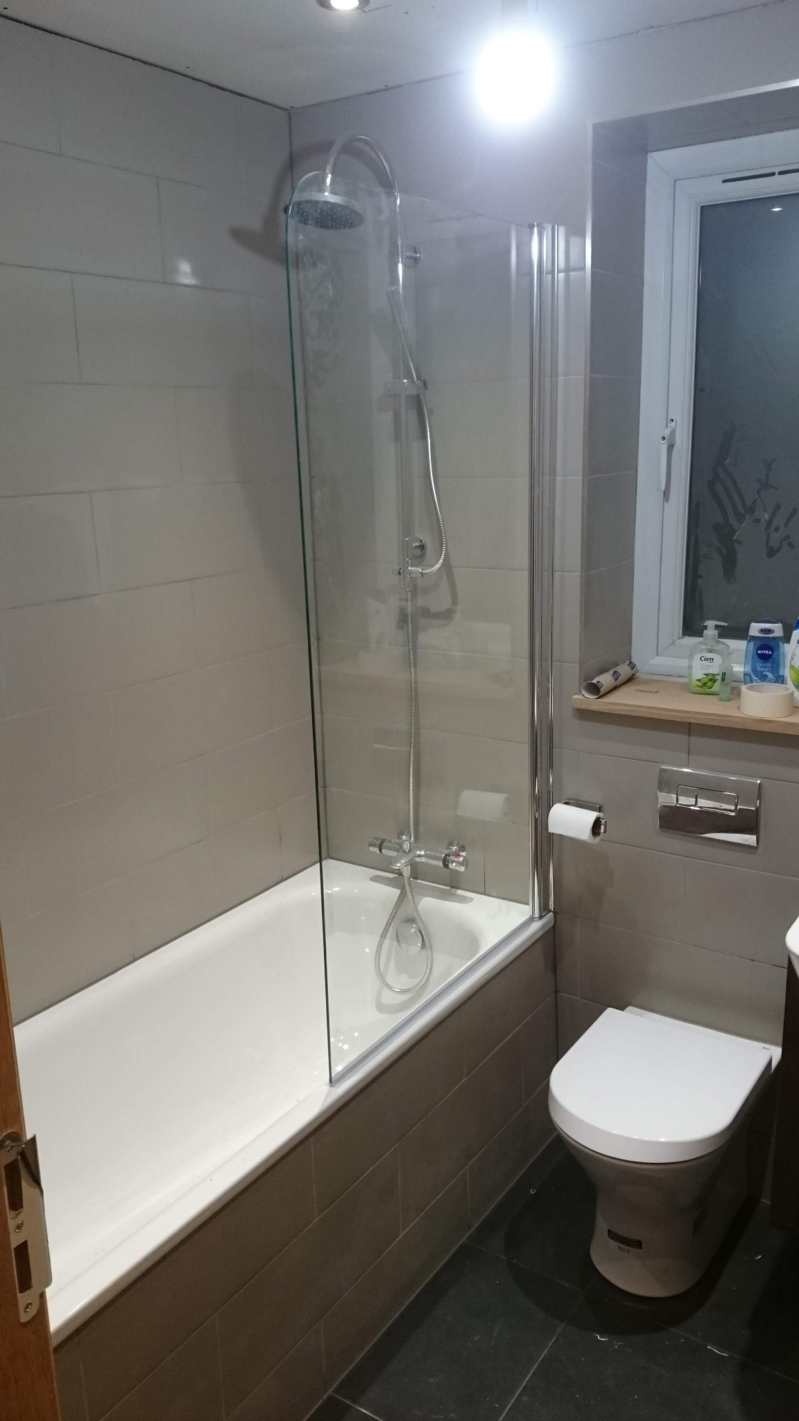 Wykończona łazienka z beżowymi płytkami, białą wanną z szklanym parawanem, toaletą i czarną podłogą, oświetlona punktowym światłem.