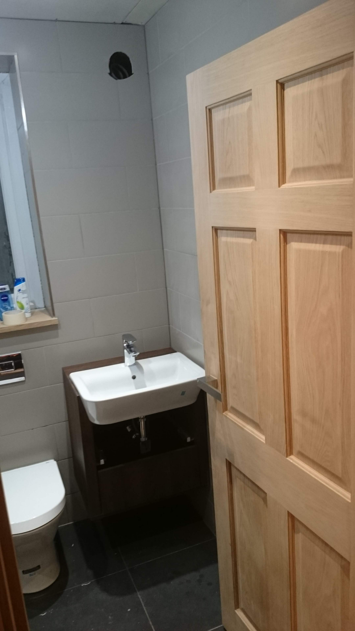 Umywalka z szafką w szarej łazience z drewnianymi drzwiami, widoczne płytki na ścianach i otwór w suficie.