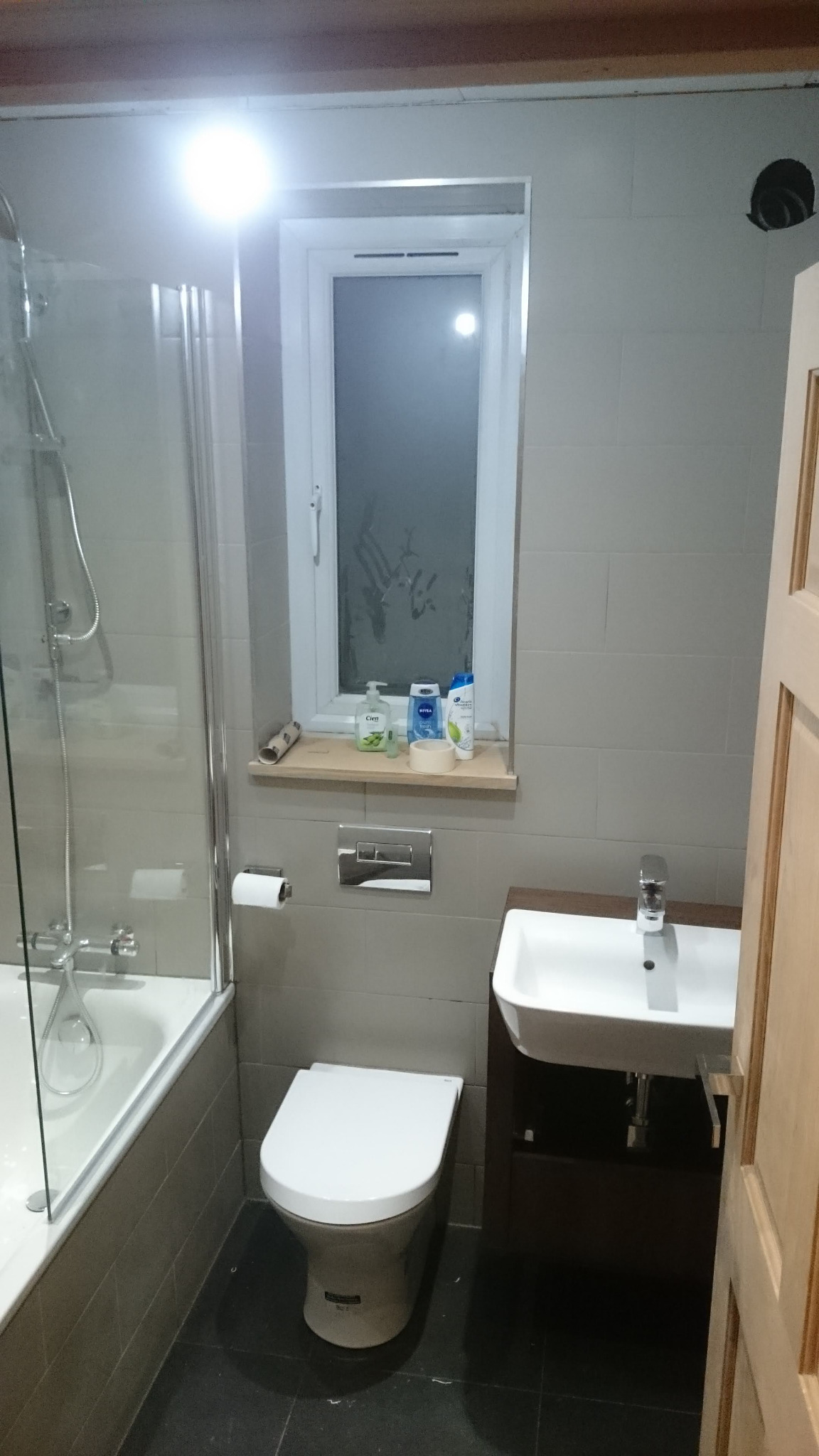 Umywalka z szafką w ciemnym odcieniu drewna, wisząca muszla klozetowa i kabina prysznicowa w łazience wykończonej szarymi płytkami, z oknem na wprost, na parapecie którego stoją kosmetyki.