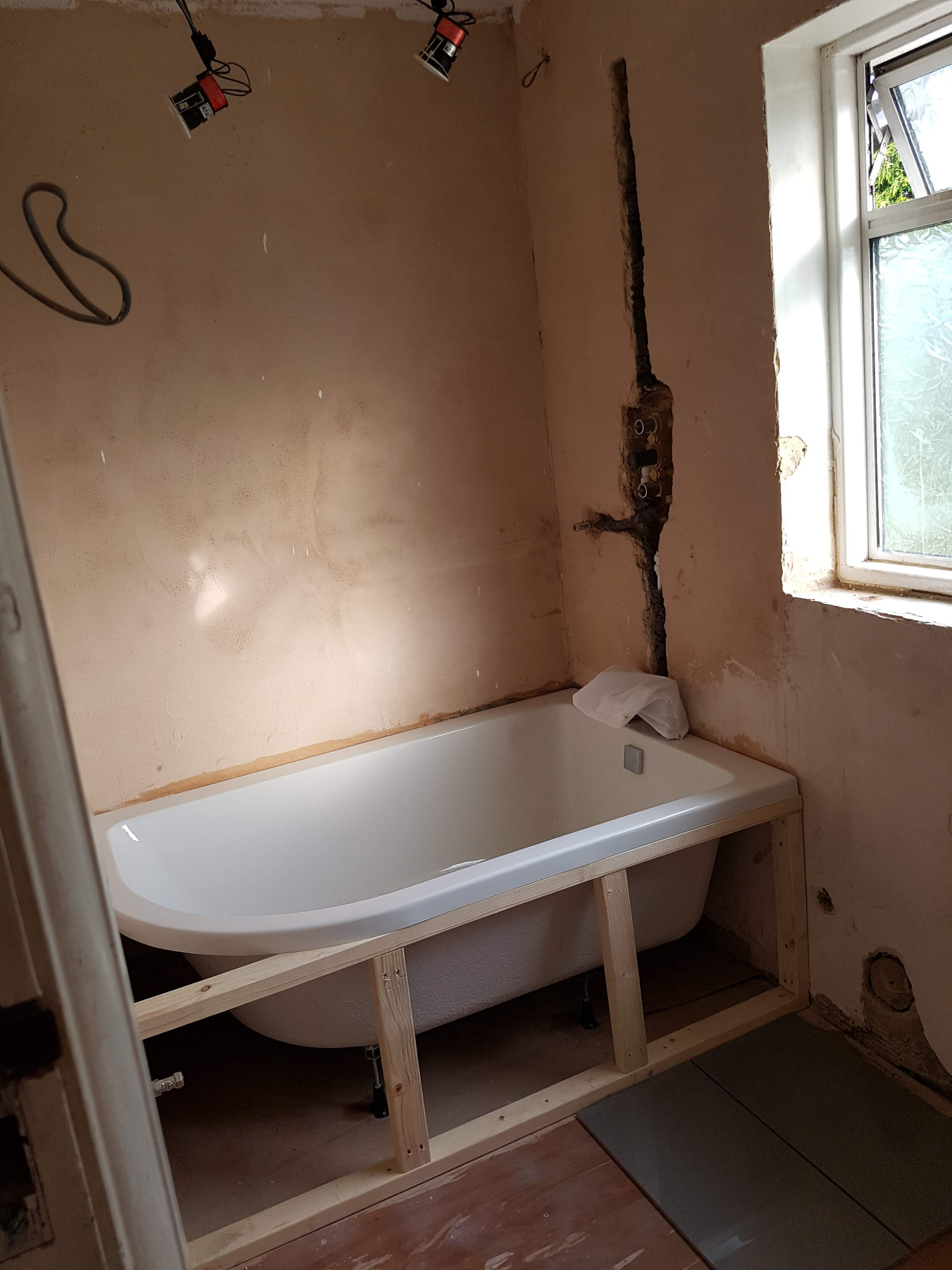 Łazienka w trakcie remontu: biała wanna osadzona w drewnianej konstrukcji, odsłonięte rury wodne w ścianie, widoczne niedoskonałości tynku i przygotowane płytki podłogowe.